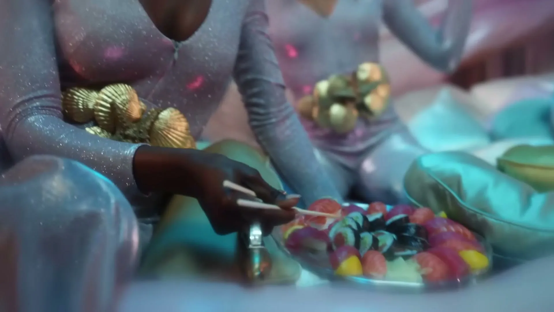 Two women in sparkly jumpsuits with gold shell accents sit on a bed, eating sushi from a platter.