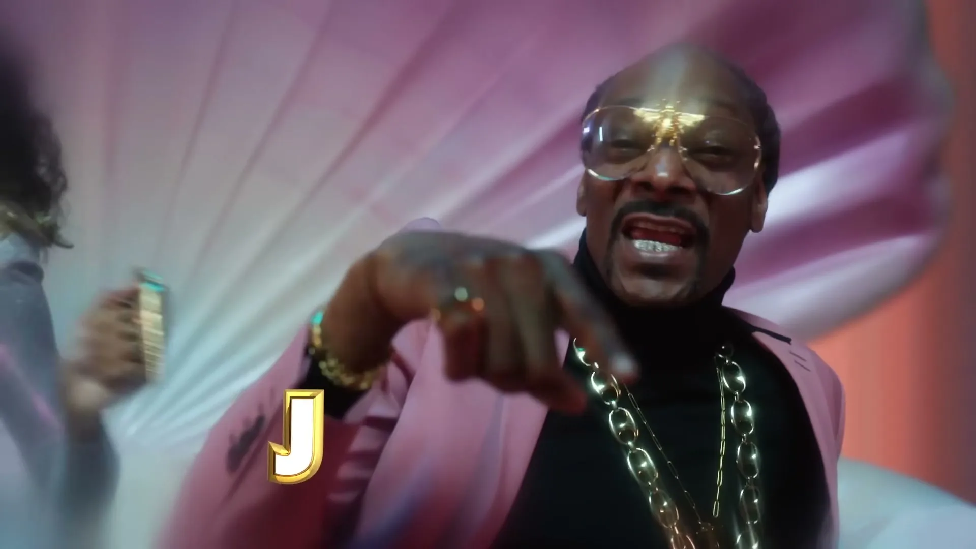 Snoop Dogg, wearing a pink suit and gold chains, points at the camera in a stylized, colorful set.