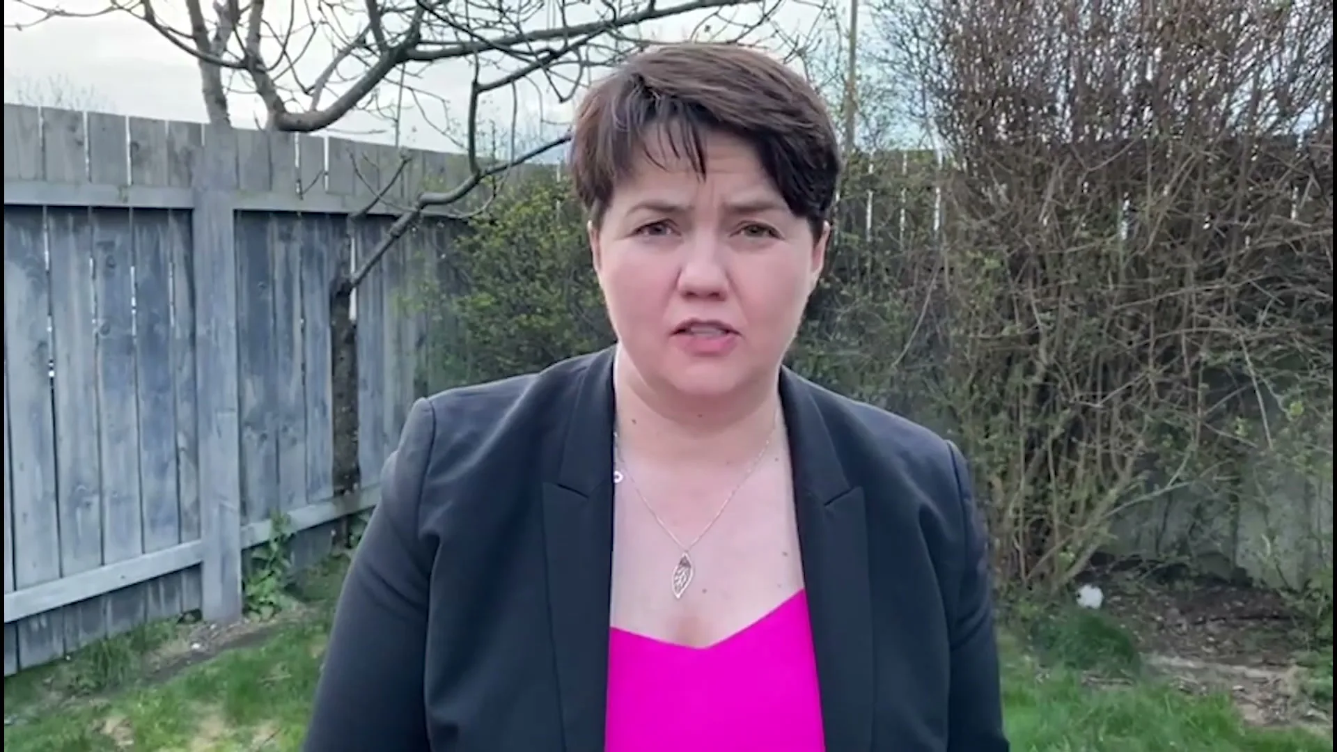 A woman with short dark hair, wearing a black blazer and a bright pink top, speaks directly to the camera in a backyard with a wooden fence.