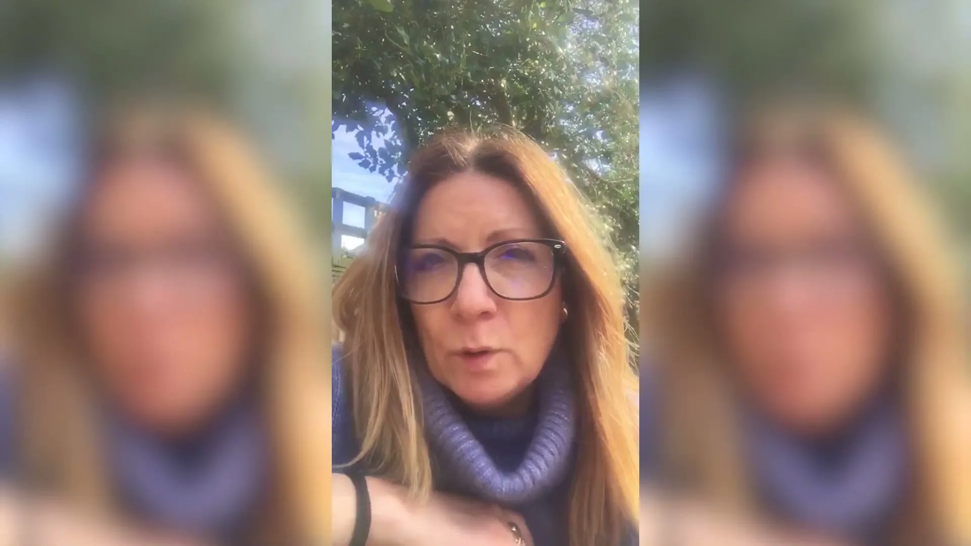 A woman with long brown hair and glasses speaks directly to the camera, wearing a blue turtleneck, outdoors in daylight.