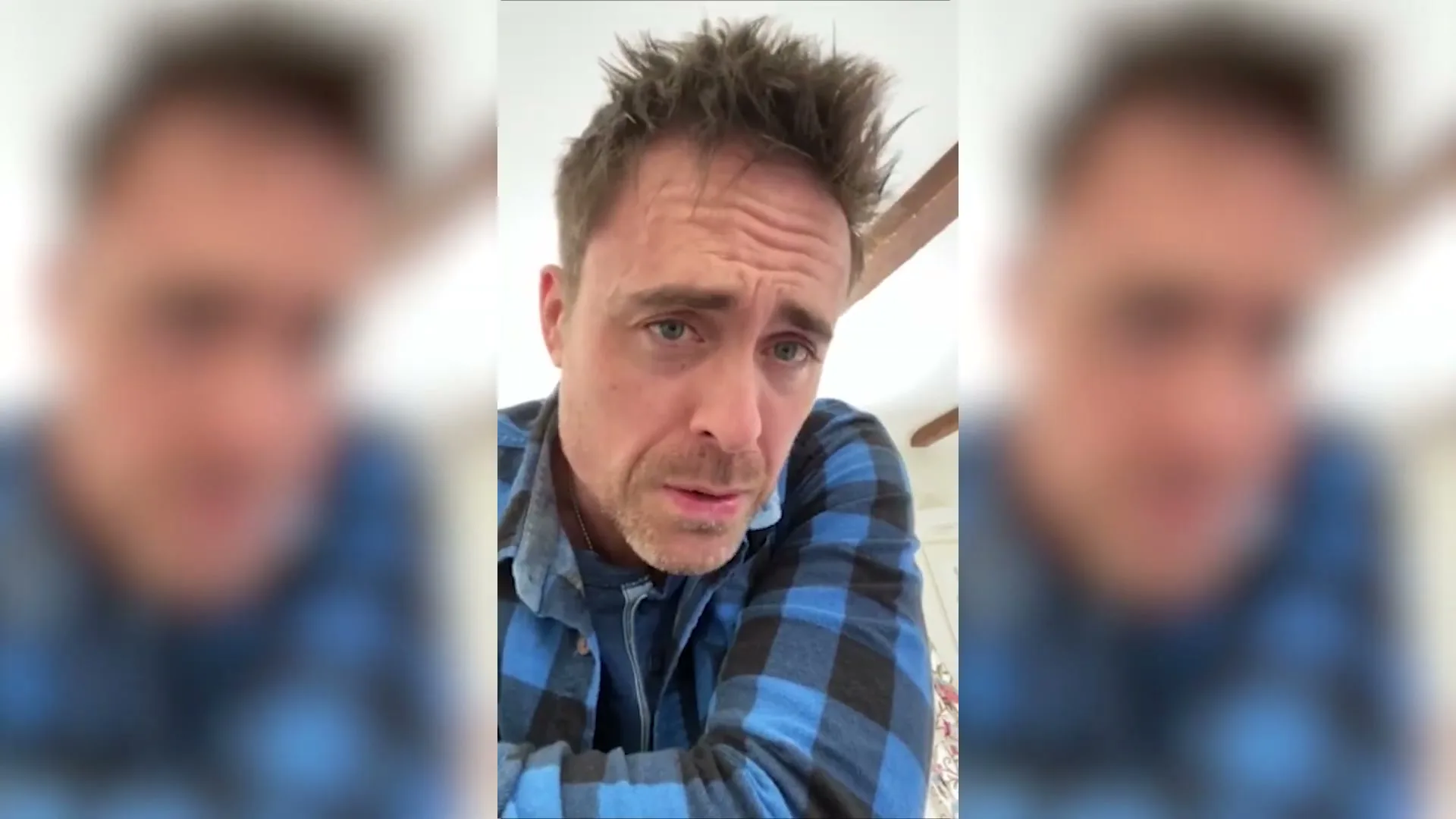 A man in his 40s, wearing a blue plaid shirt, looks directly at the camera with a concerned expression in an interior setting.