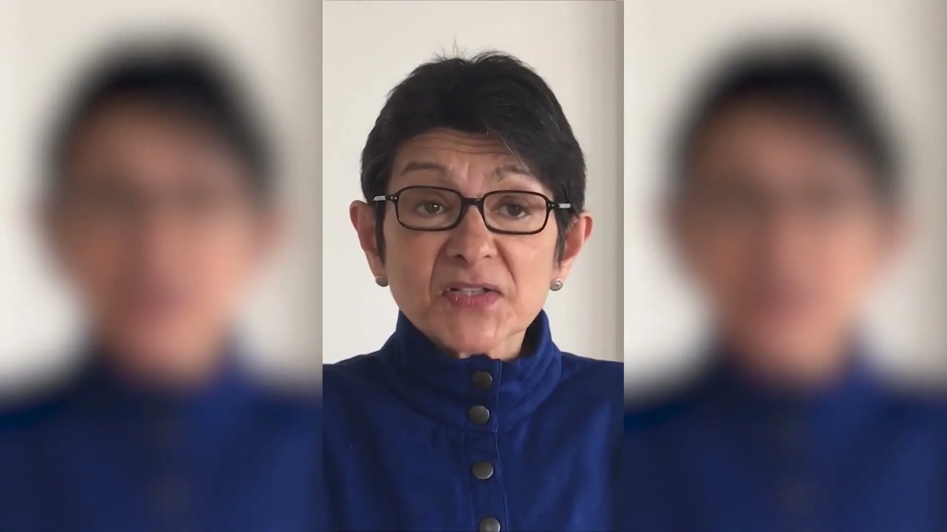 A woman with short dark hair and glasses, wearing a blue jacket, speaks directly to the camera against a plain white background.