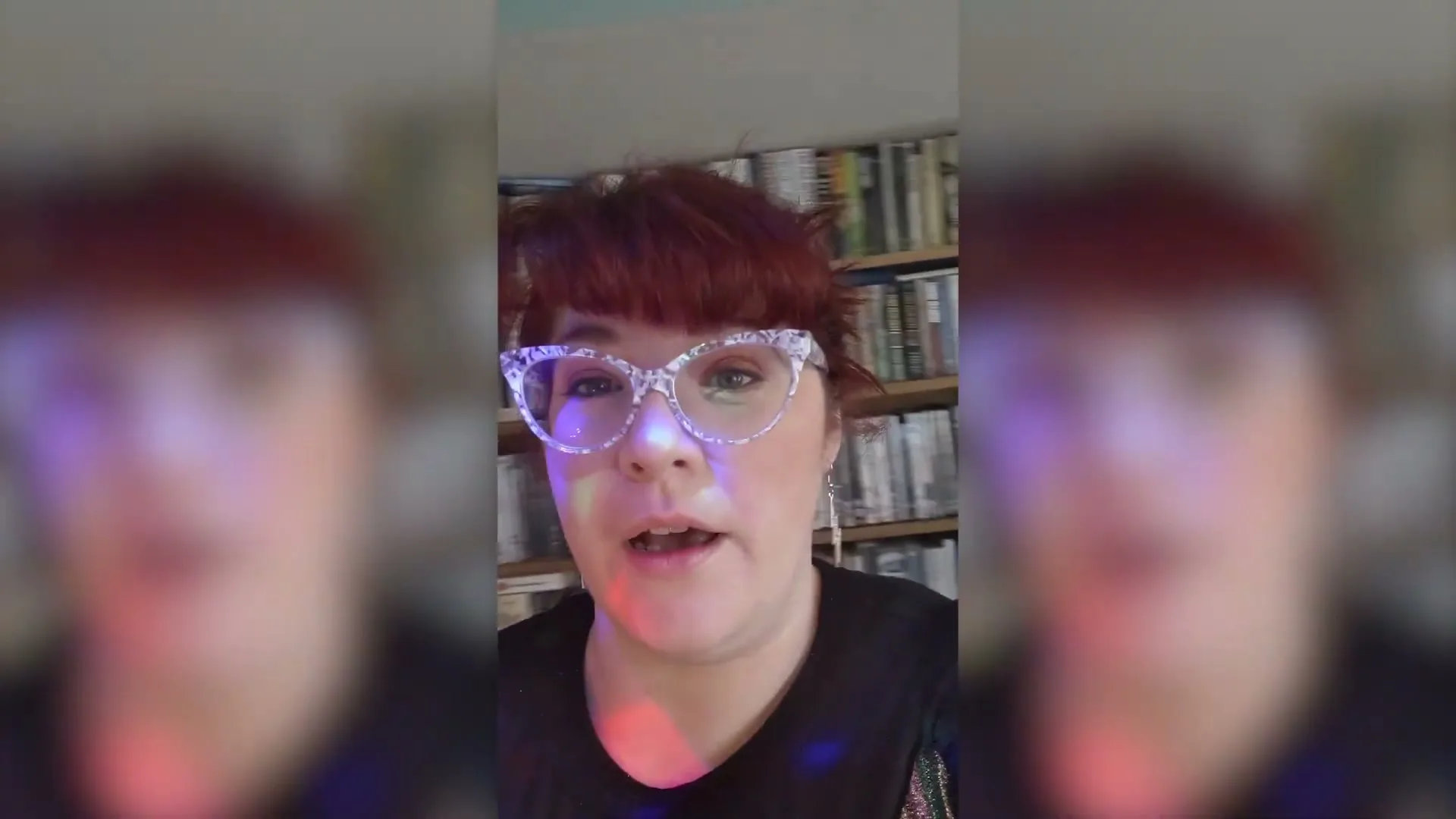 A woman with red hair and glasses talks to the camera, with a bookshelf visible in the background, as disco lights flash around her.