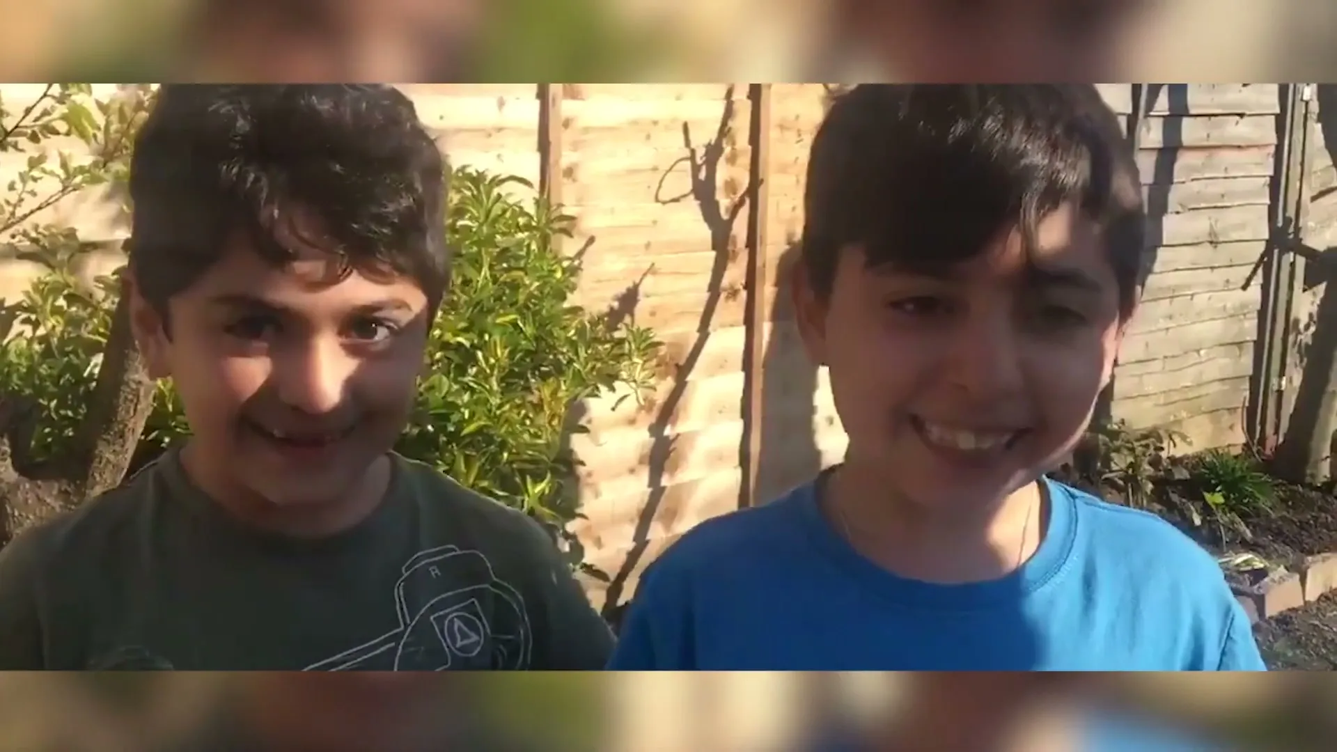 Two young boys, one in a green t-shirt and one in a blue t-shirt, stand side-by-side in a sunny backyard, smiling at the camera.