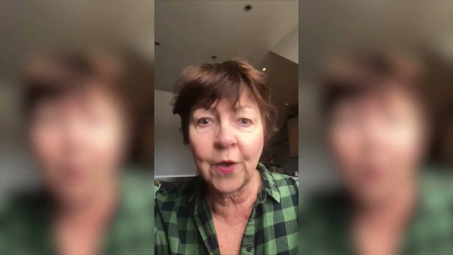A woman with short brown hair and a green plaid shirt speaks directly to the camera in a medium closeup shot, indoors.