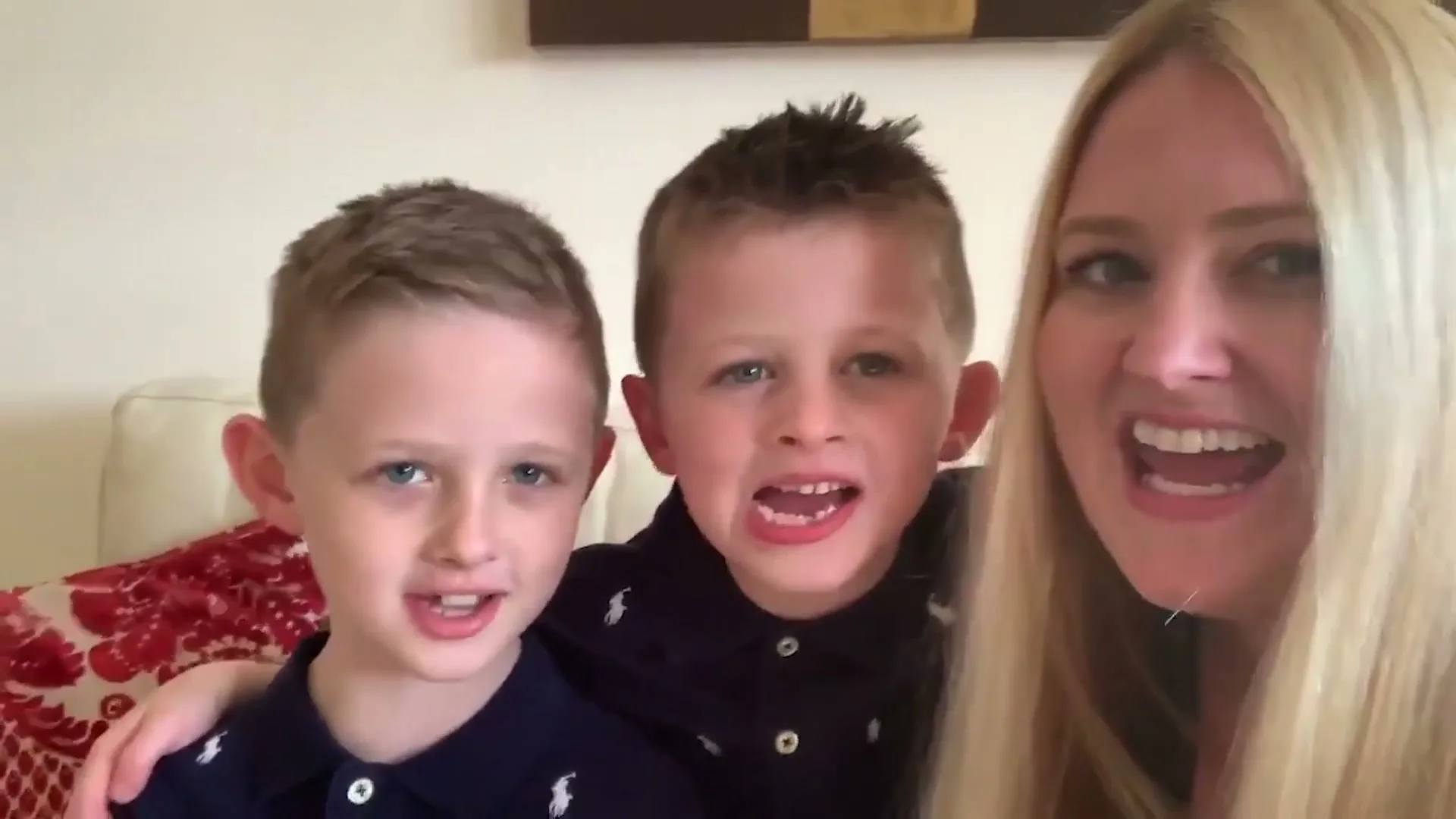 A blonde woman and two young boys in polo shirts are seen in a medium shot, smiling and talking directly to the camera in a living room.