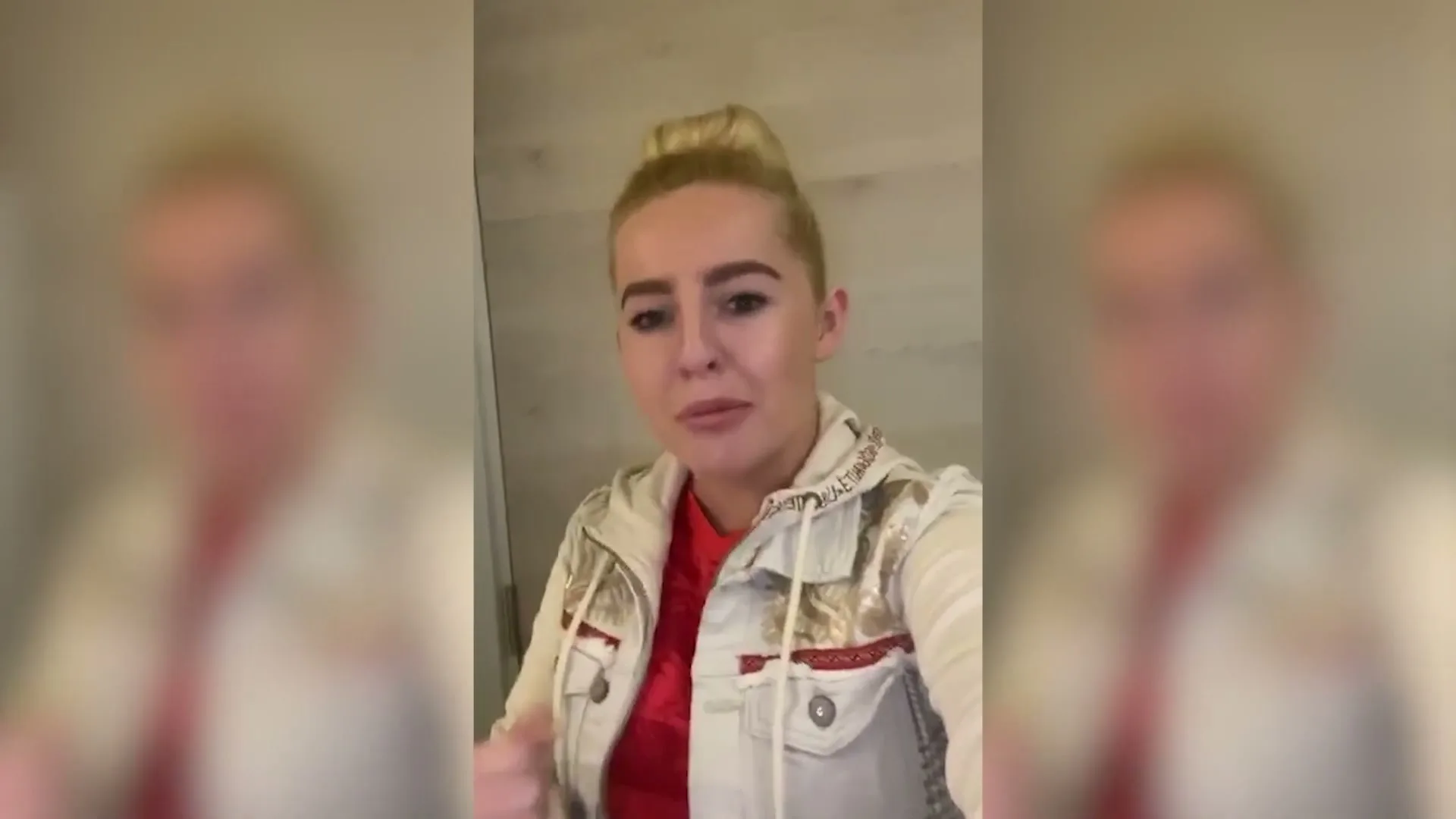 A young woman with blonde hair in a bun, wearing a red top and a light jacket, speaks directly to the camera in an interior setting.