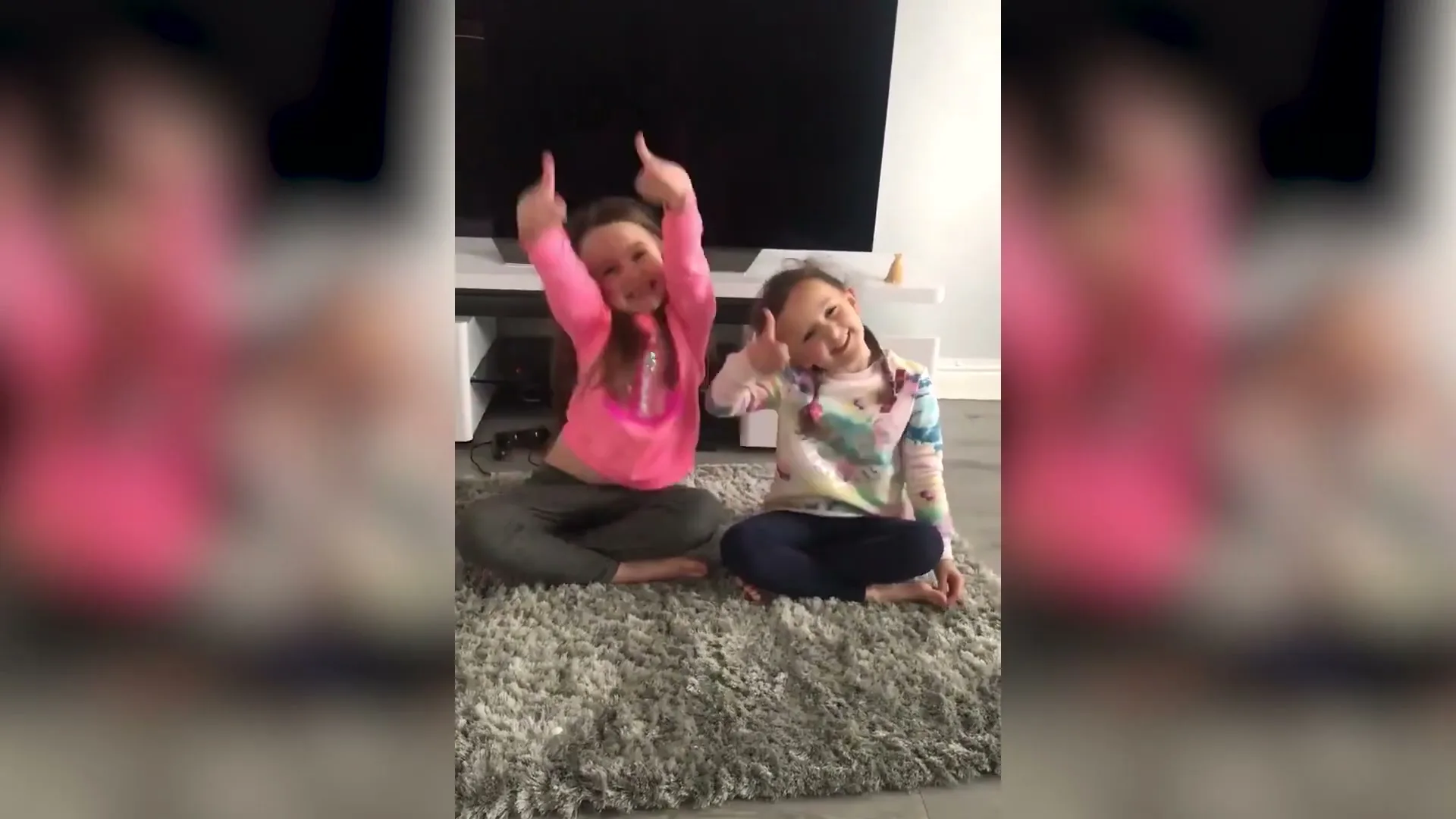 Two young girls sit cross-legged on a rug in a living room, smiling and giving thumbs up to the camera.