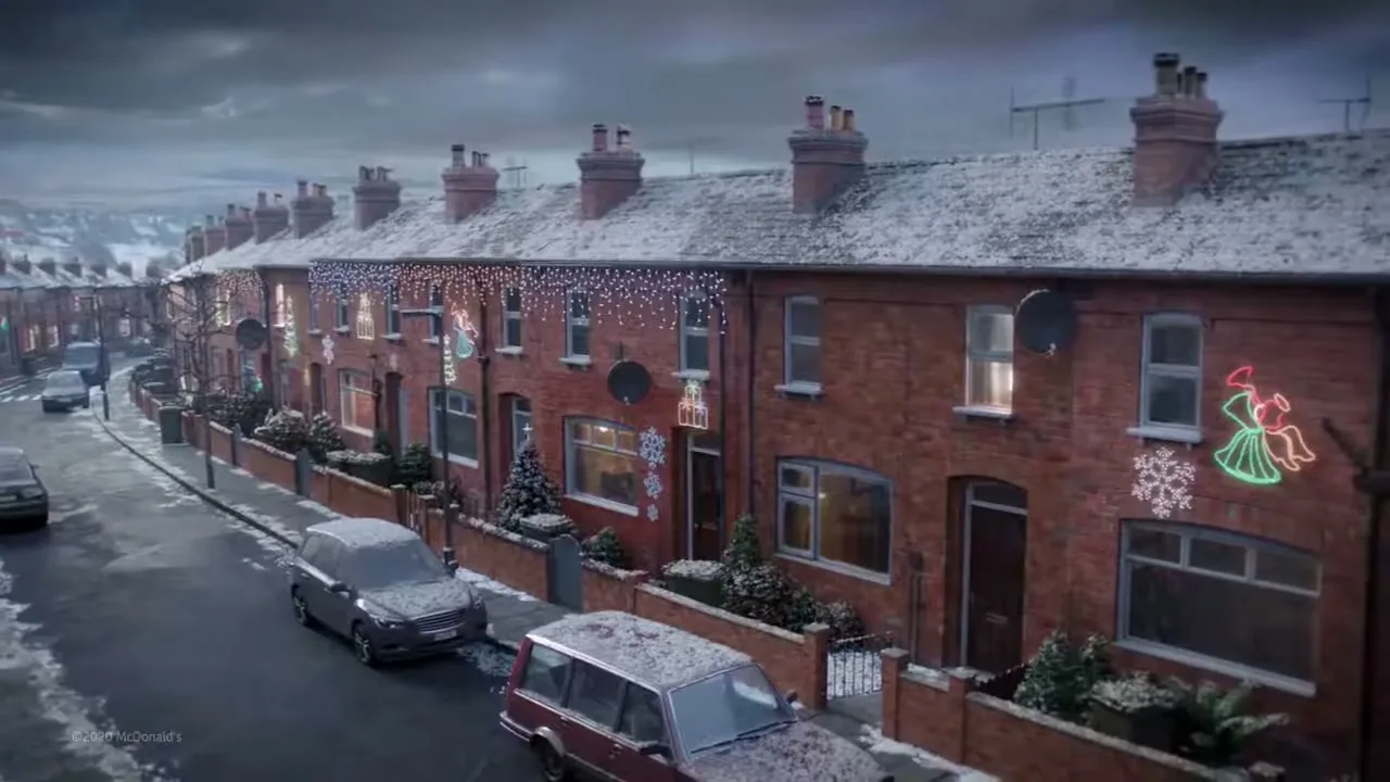 A wide shot of a snowy street with brick houses decorated with Christmas lights under a cloudy sky.