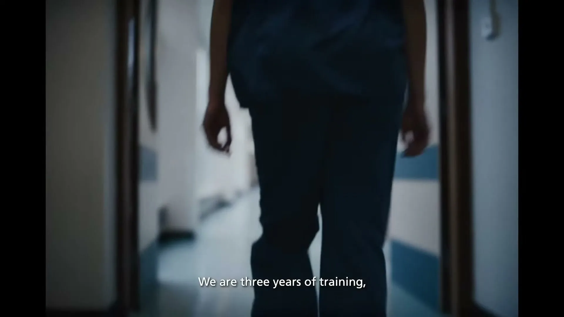 A person in blue scrubs walks away from the camera down a hospital corridor, with natural light streaming in from windows on the left.