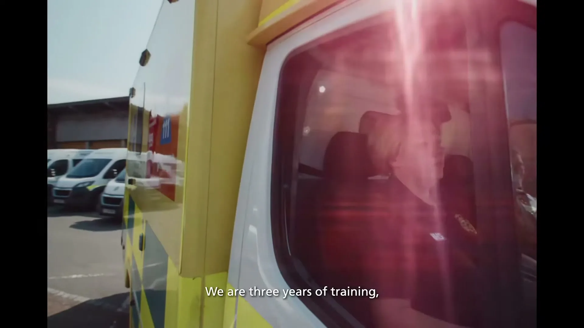 A female paramedic in a dark uniform with an NHS logo sits in the driver's seat of a yellow ambulance, looking forward, with other ambulances parked in the background.
