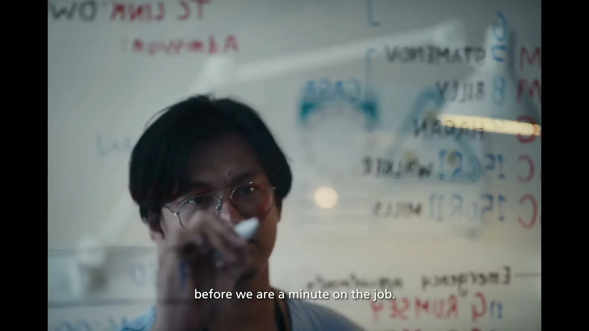 A male healthcare worker in glasses and scrubs writes on a clear board with a marker, seen through the board.