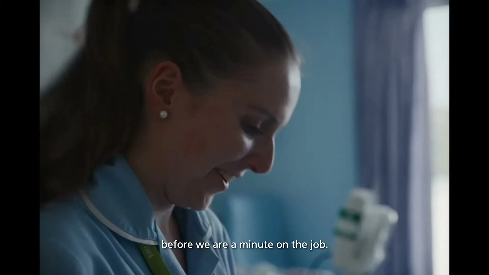 A young female nurse in blue scrubs smiles while looking down, with medical equipment visible in the background of a hospital room.