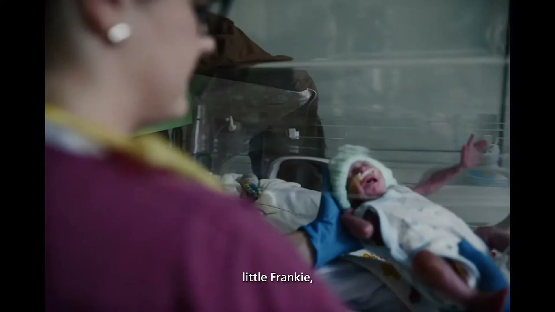 A nurse in scrubs and blue gloves holds a tiny, crying premature baby in a hospital incubator.