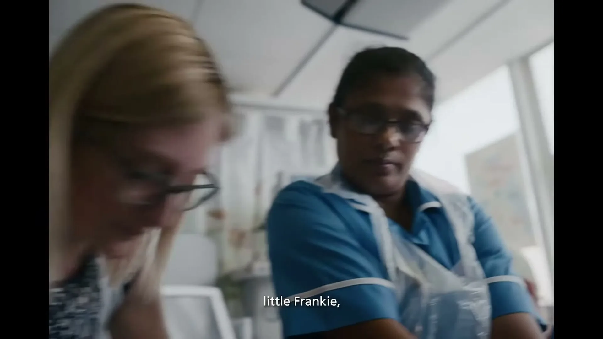 Two female nurses, one with blonde hair and glasses, the other with dark hair and glasses, are seen in a hospital room, looking down and working.