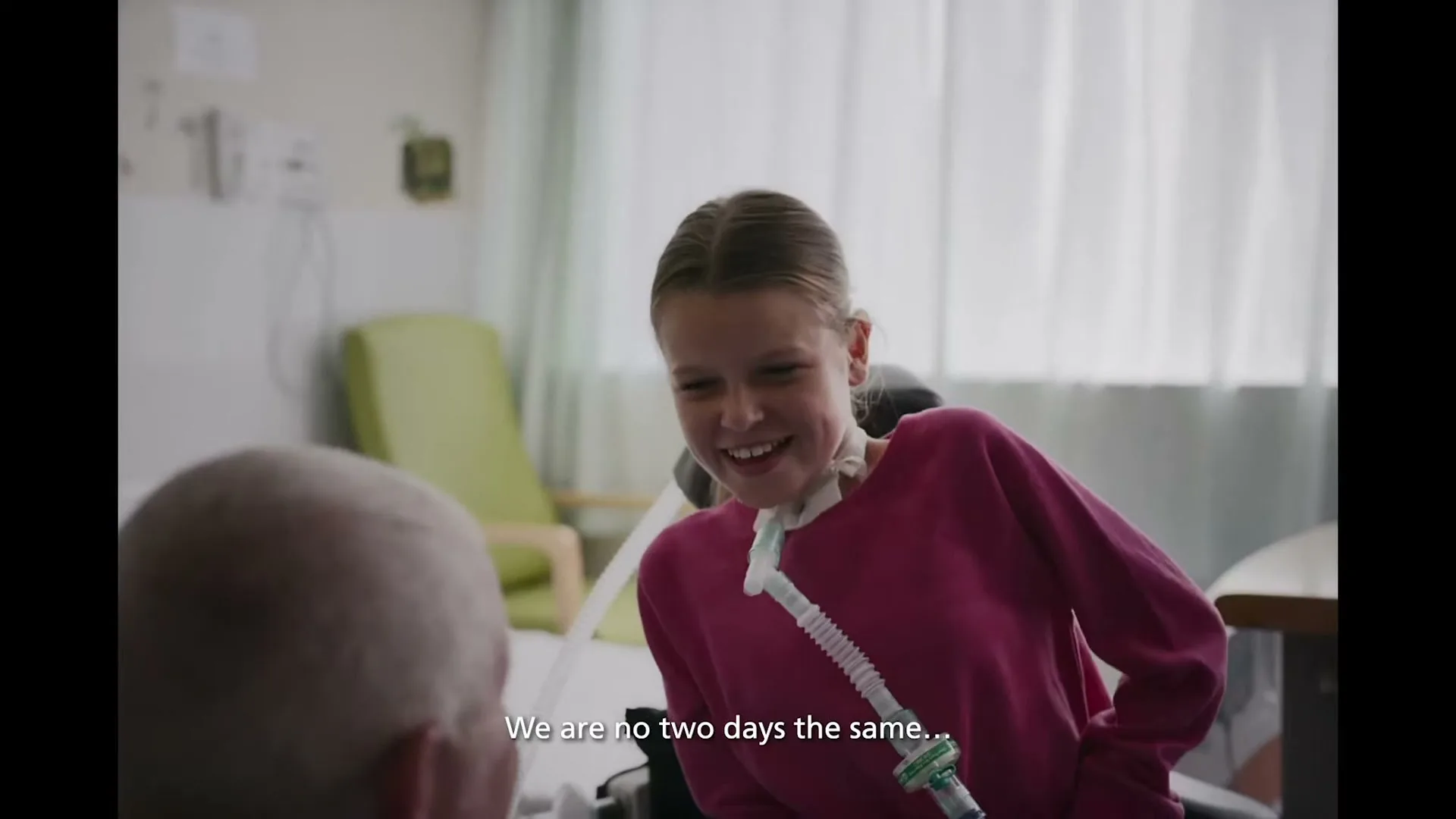 A young female patient with a tracheostomy tube smiles at an unseen person in a hospital room.