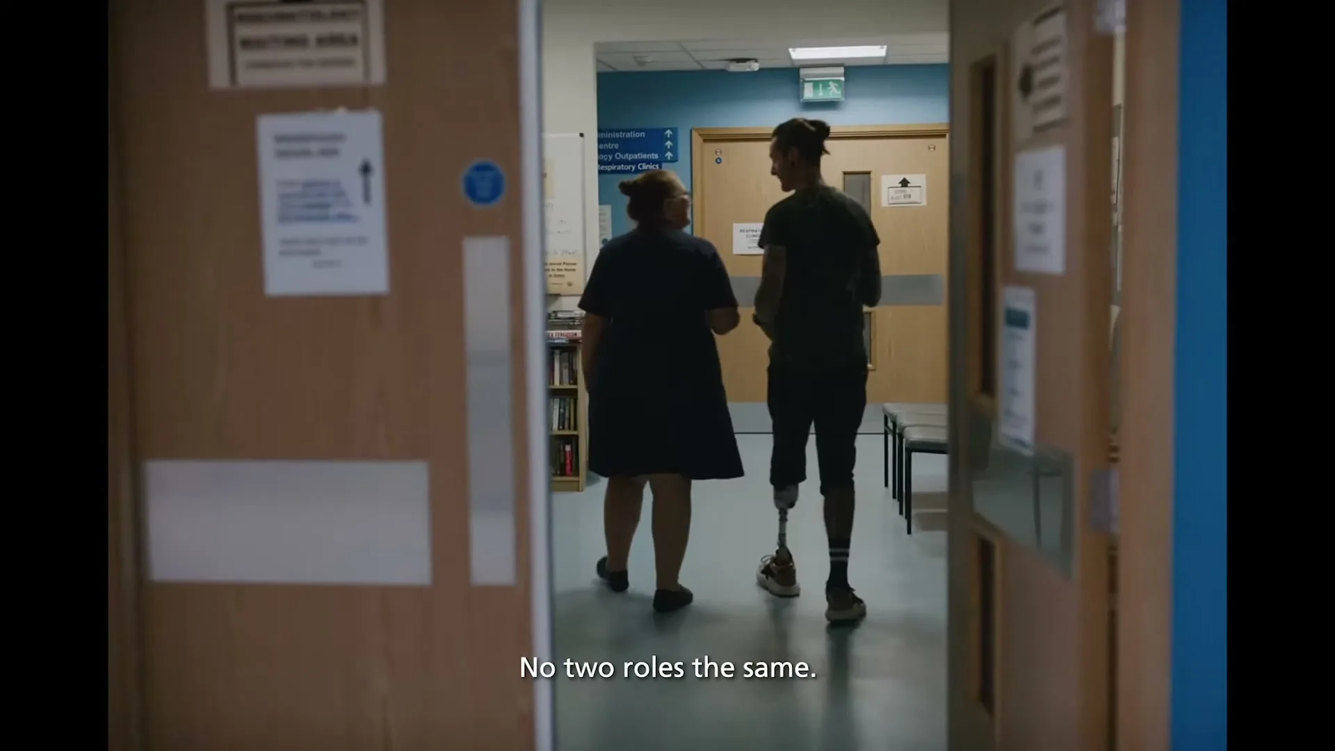 A female nurse and a male patient with a prosthetic leg walk down a hospital corridor, seen from behind through a doorway.