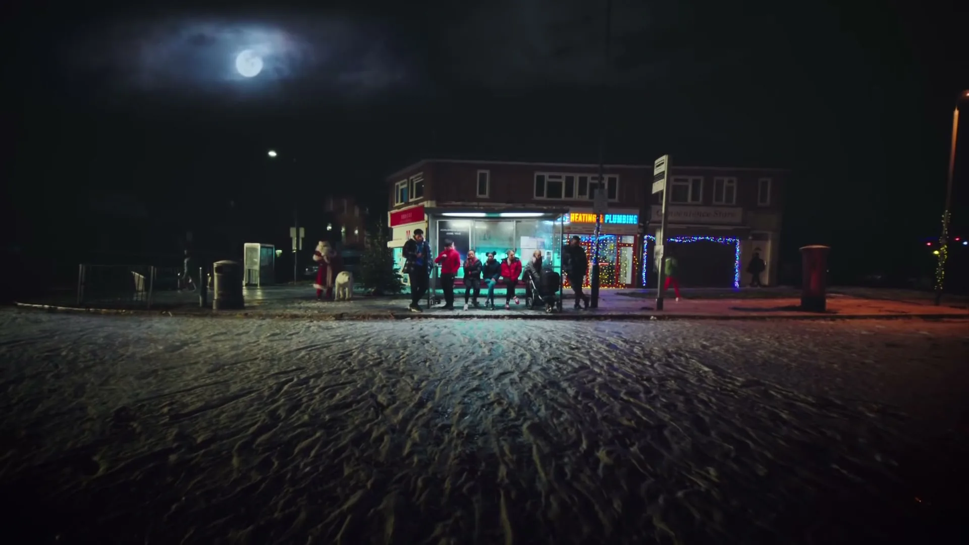 A group of diverse people in winter clothes stand at a bus stop in front of a small building with neon signs at night under a full moon.