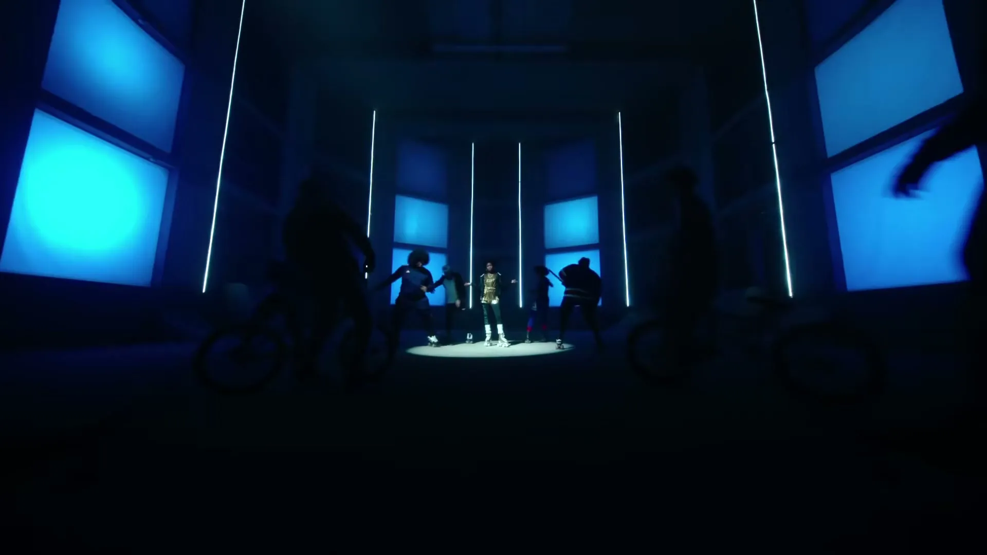 A young man in a patterned shirt stands in a spotlight as other people on bikes ride past him in a dark, blue-lit room.