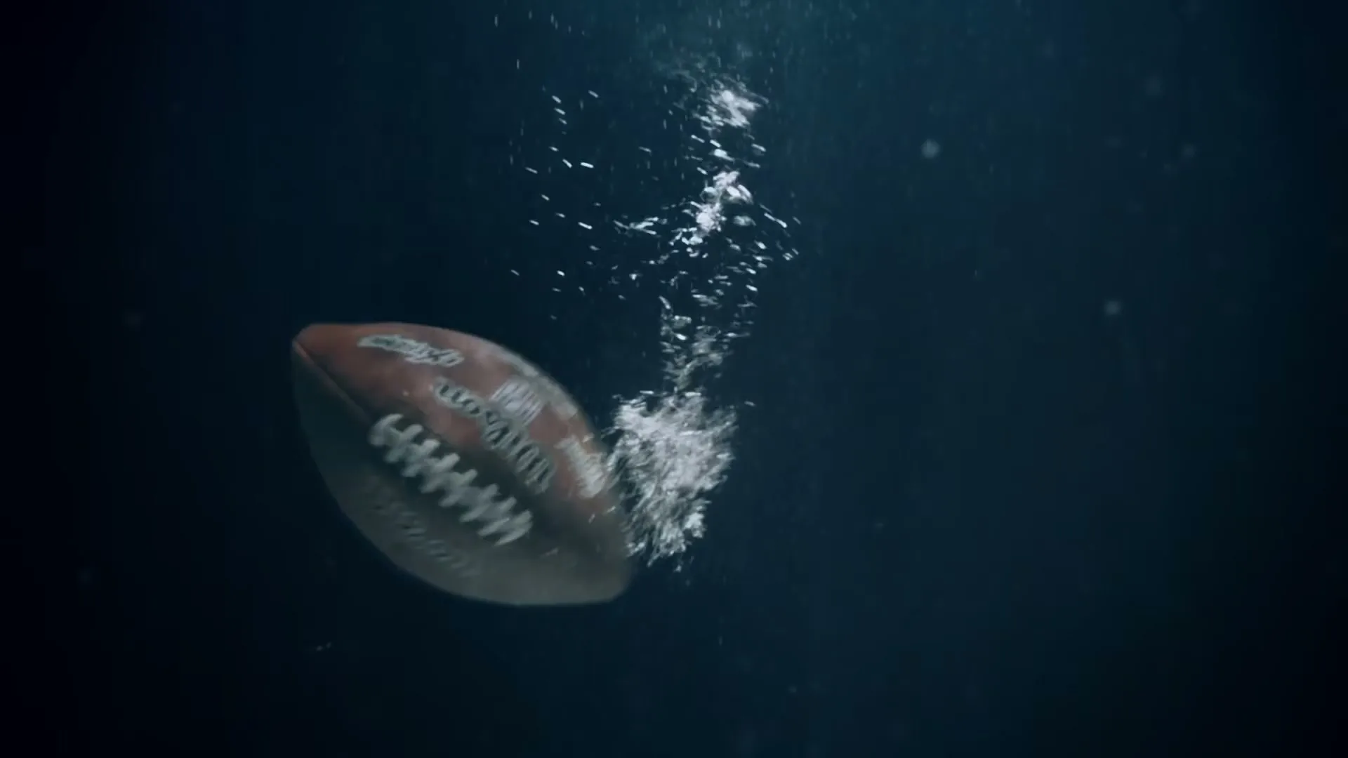 A football is thrown into water, creating splashes and bubbles as it sinks into the dark blue depths.