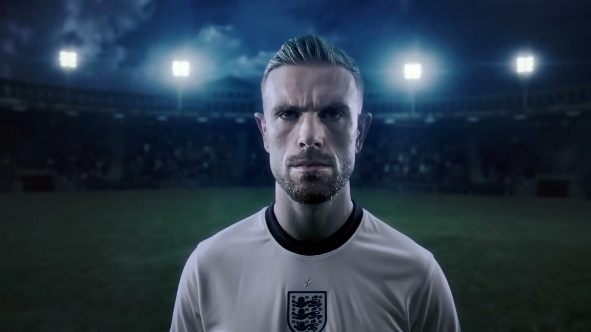 A male soccer player in a white jersey stands on a stadium field at night, looking directly at the camera with a serious expression.