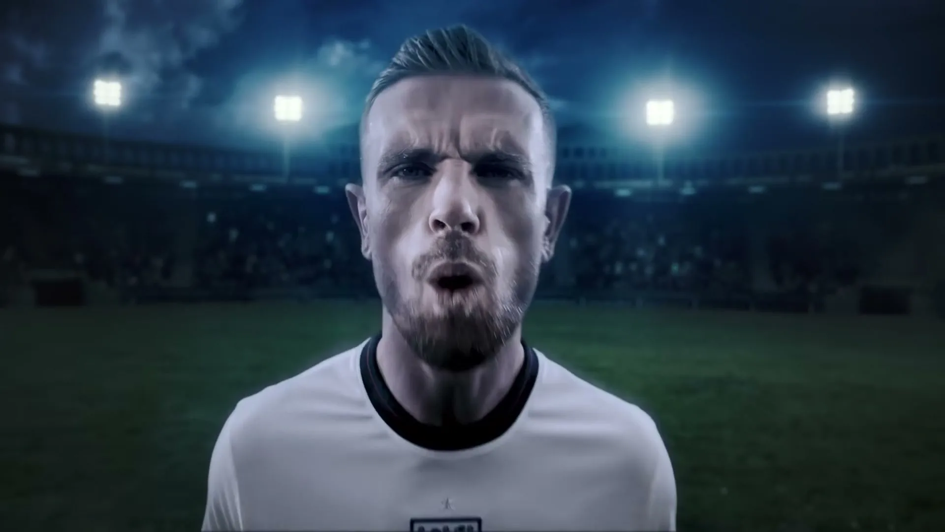 A male soccer player in a white jersey looks intensely at the camera on a stadium field at night, then quickly turns his head.