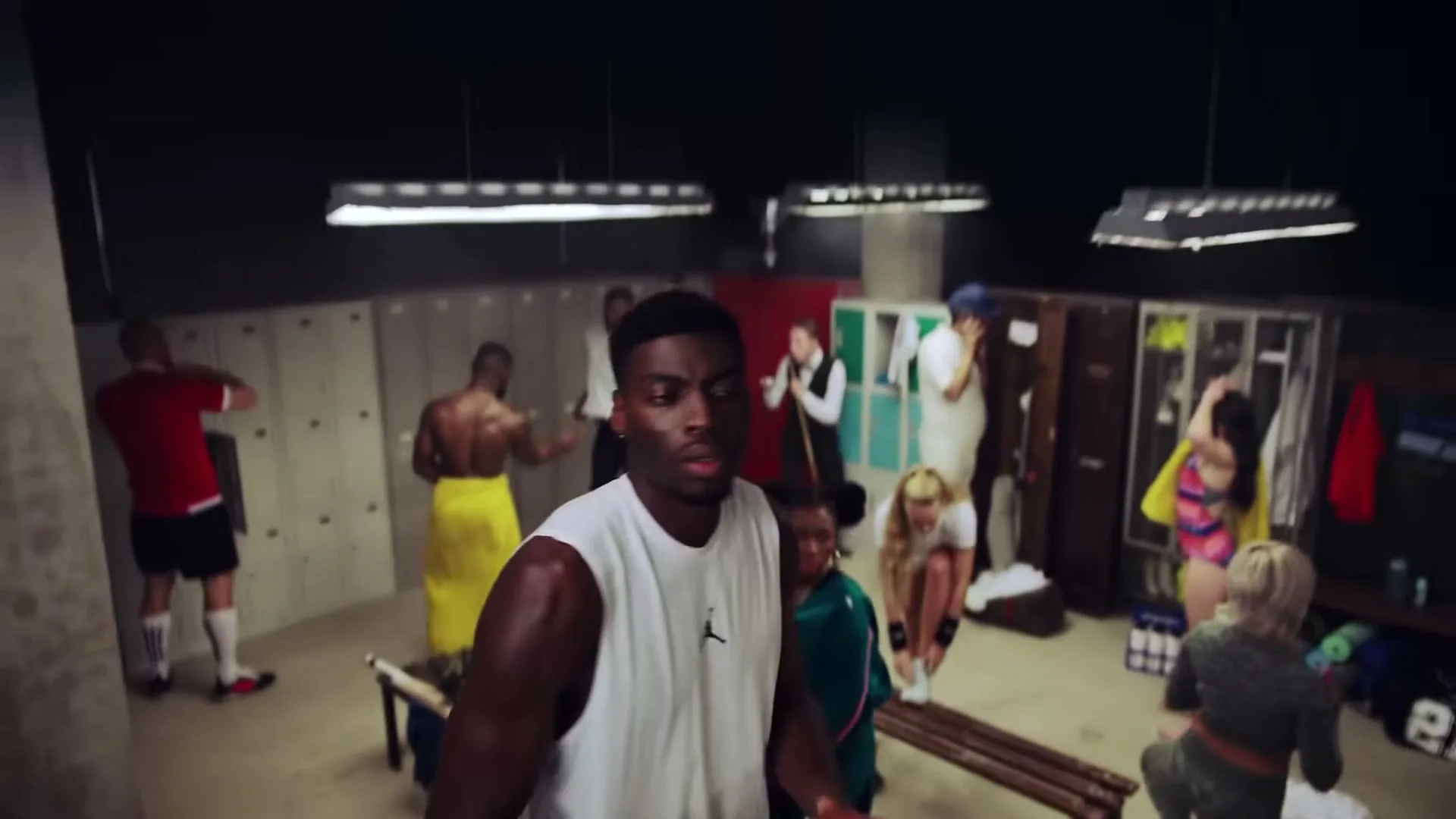 A young Black man in a white tank top dribbles a basketball in a busy locker room with other athletes getting ready.