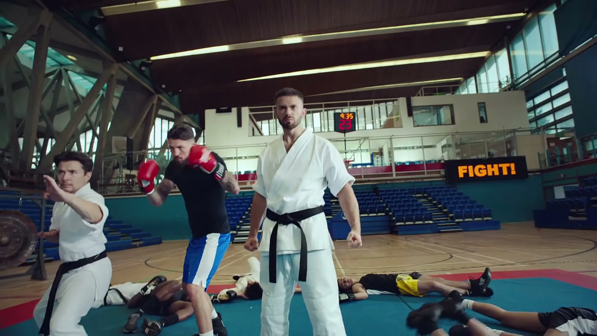 A male karateka in a white gi and black belt stands confidently in a gym, surrounded by other athletes in various states of action or repose.