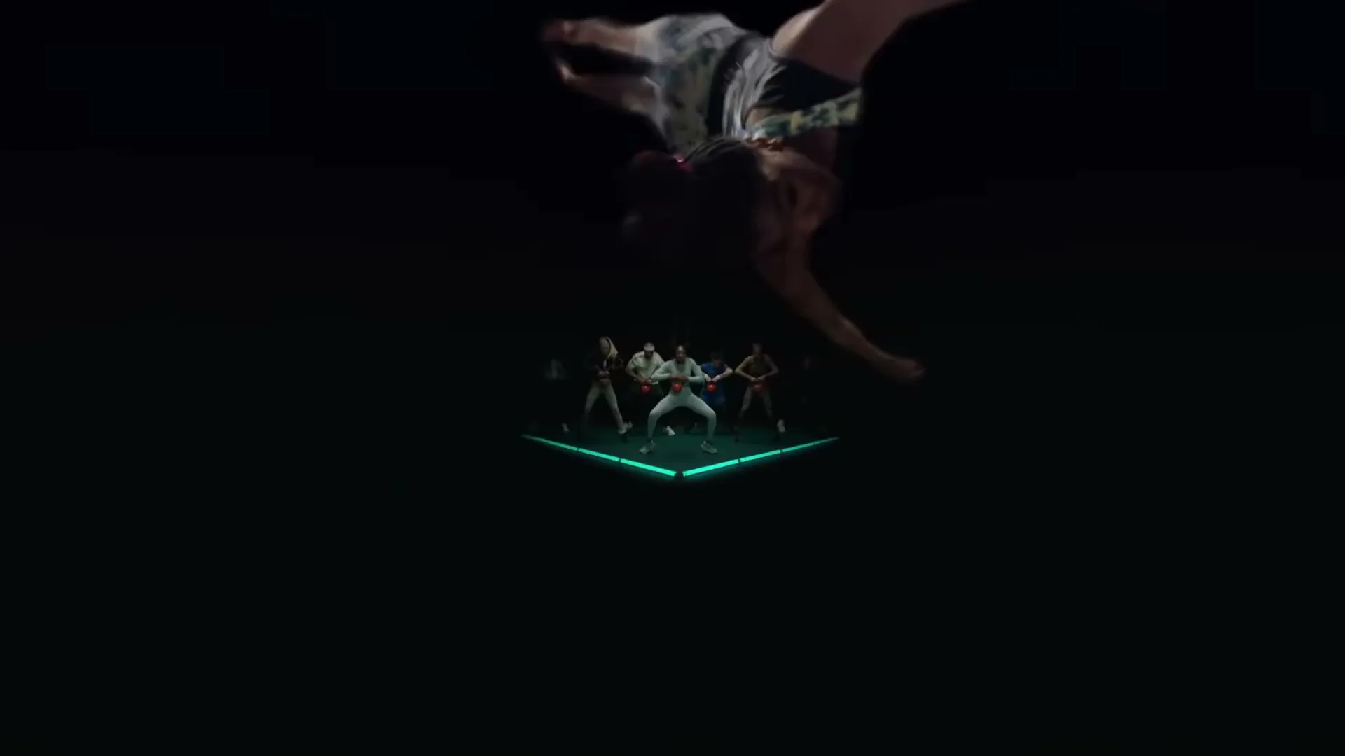 A group of people in athletic wear dance on a glowing green platform in a dark studio, while a person swings a long white pole.