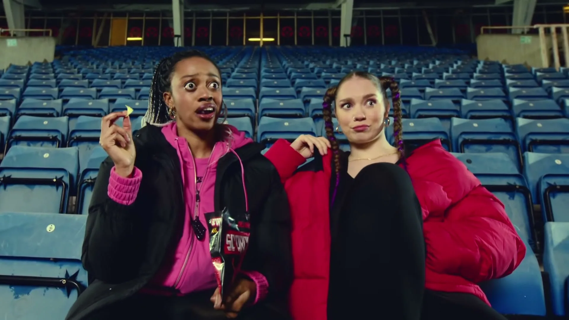 Two young women in puffer jackets sit in empty stadium seats, one holding a snack and looking surprised, the other looking unimpressed.