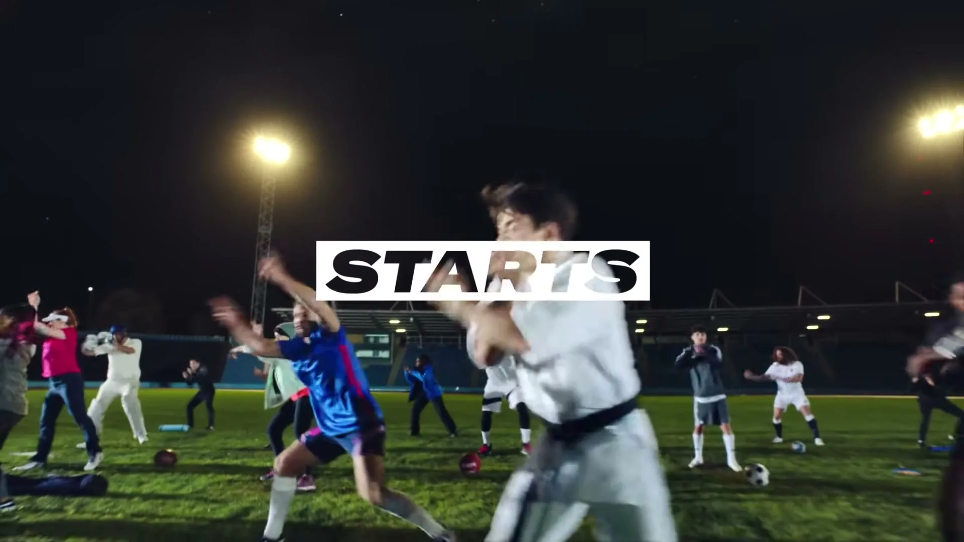 A diverse group of athletes in various sports uniforms performs dynamic exercises on a floodlit grass field at night, with "STARTS" text appearing over them.