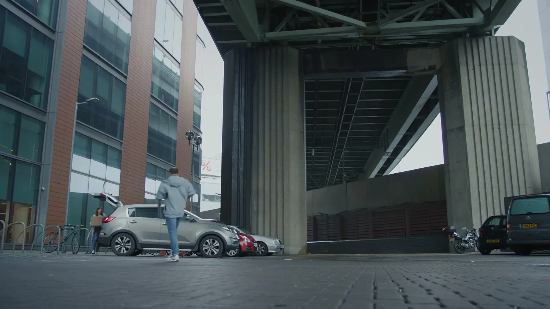A young man in a grey hoodie and jeans walks towards a silver SUV parked under a bridge, while a woman stands by the open trunk.