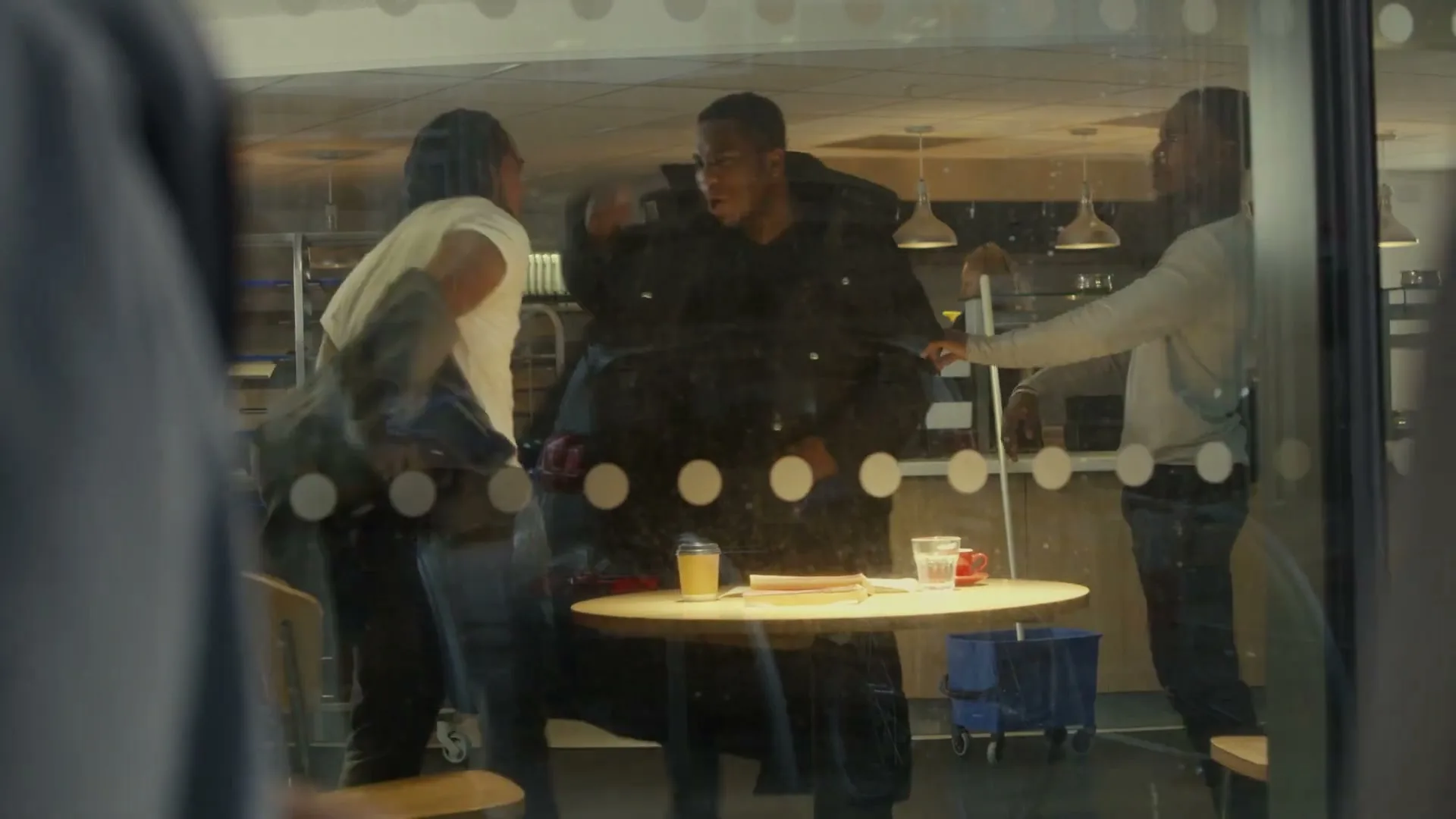 Three young men are having a heated conversation in a cafe, which escalates into a physical altercation, viewed through a window.
