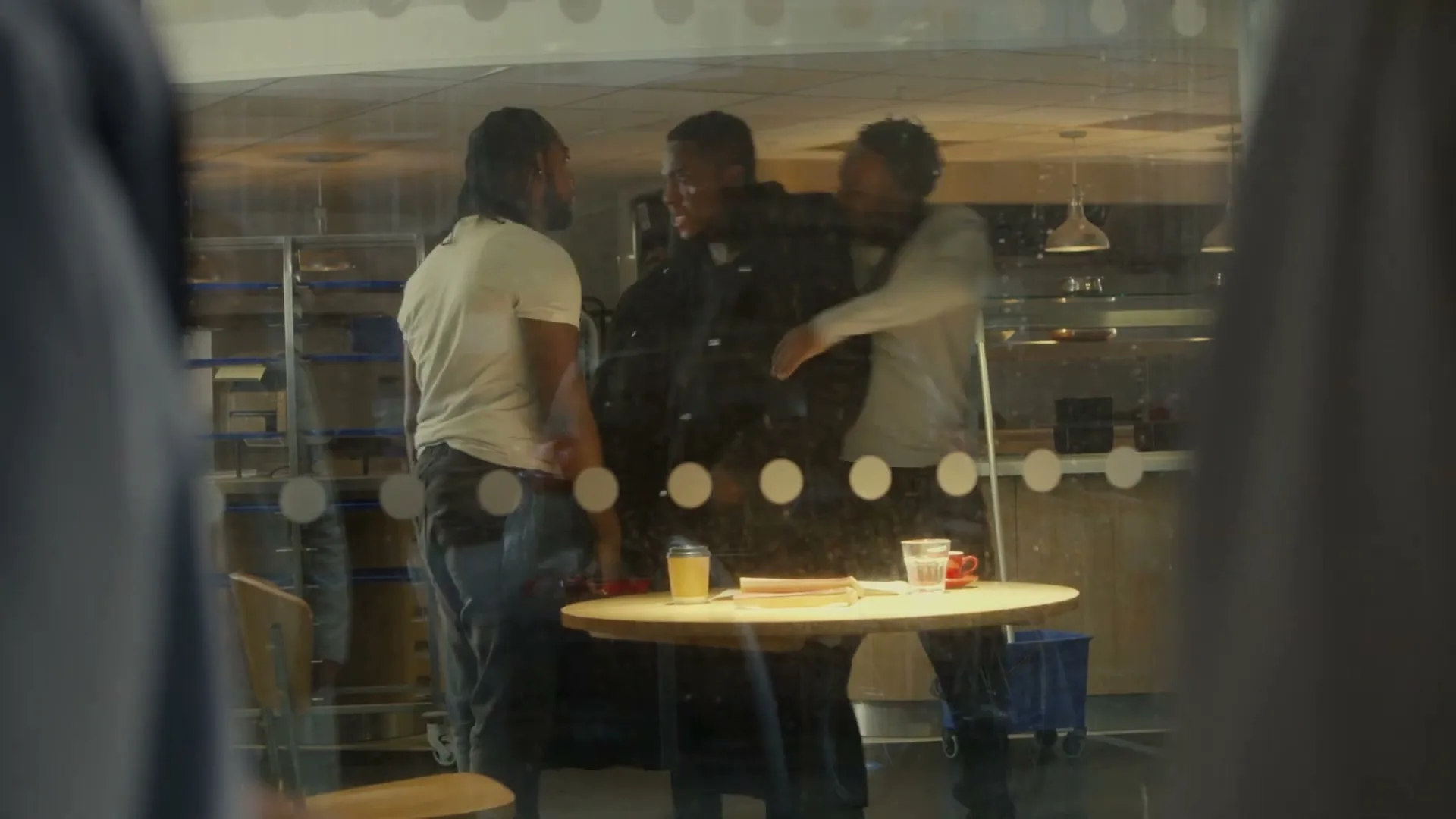Three young men are seen through a glass partition, two of them holding back a third man who appears agitated, in a modern interior space.