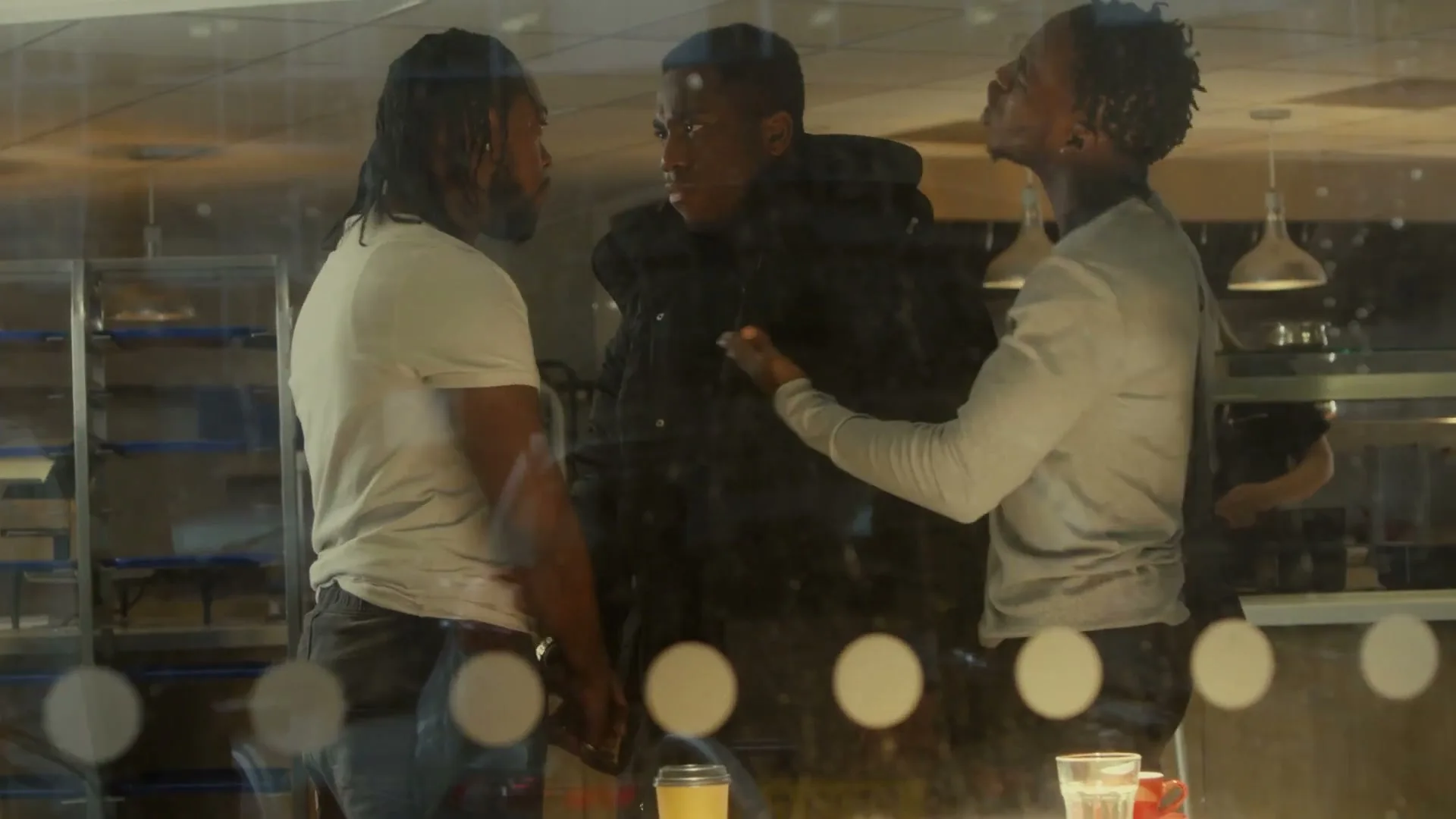 Three young men, two with dreadlocks, stand around a table in a cafe, engaged in a heated conversation.