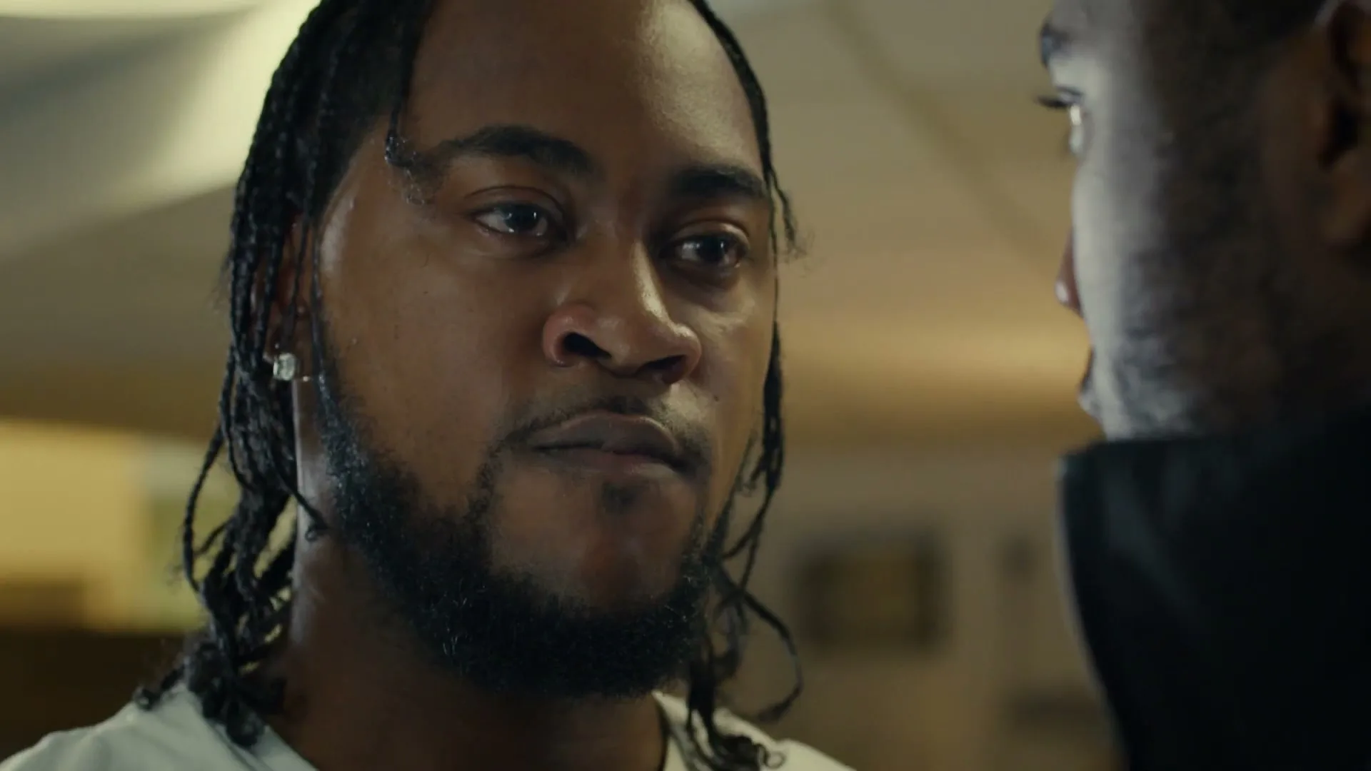 A Black man with braids and a beard looks intently at another person, whose back is partially visible, in an interior setting.