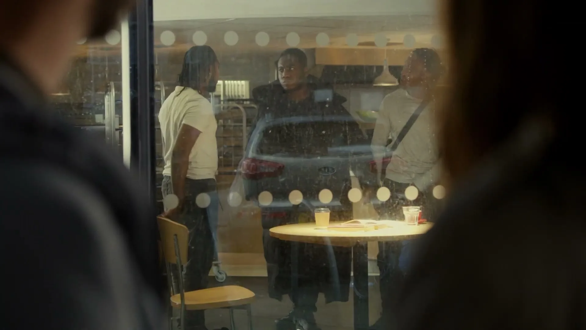 Three young Black men stand outside a cafe, seen through a window with reflections, while two blurred figures observe them from inside.