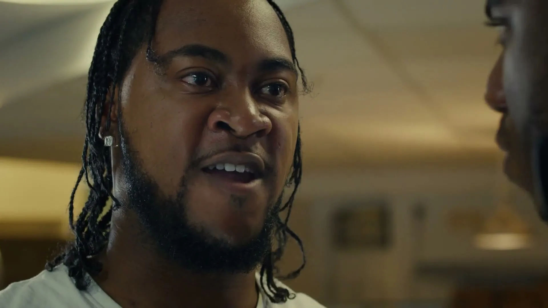 A Black man with braided hair and a beard yells intensely at another person, his face contorted in anger, in an interior setting.