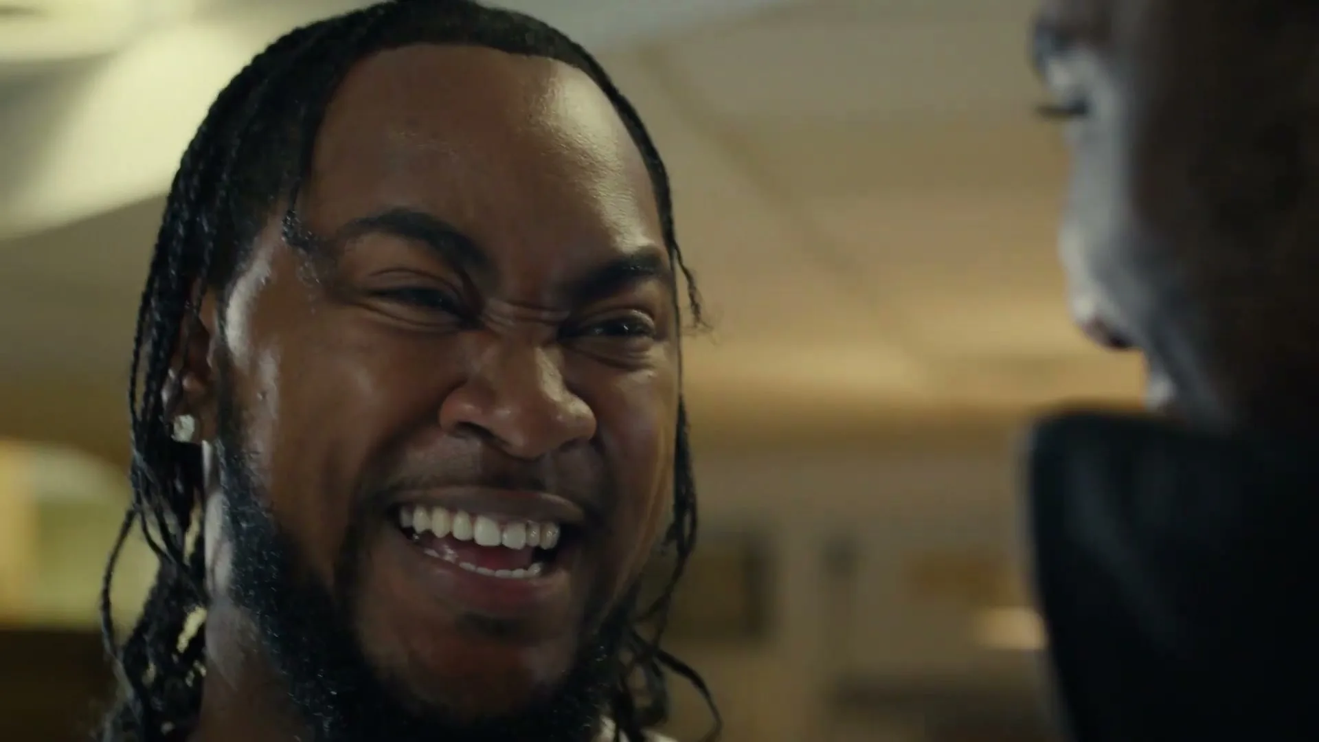 A Black man with braided hair and a beard looks at another person, then laughs heartily, in an interior setting.