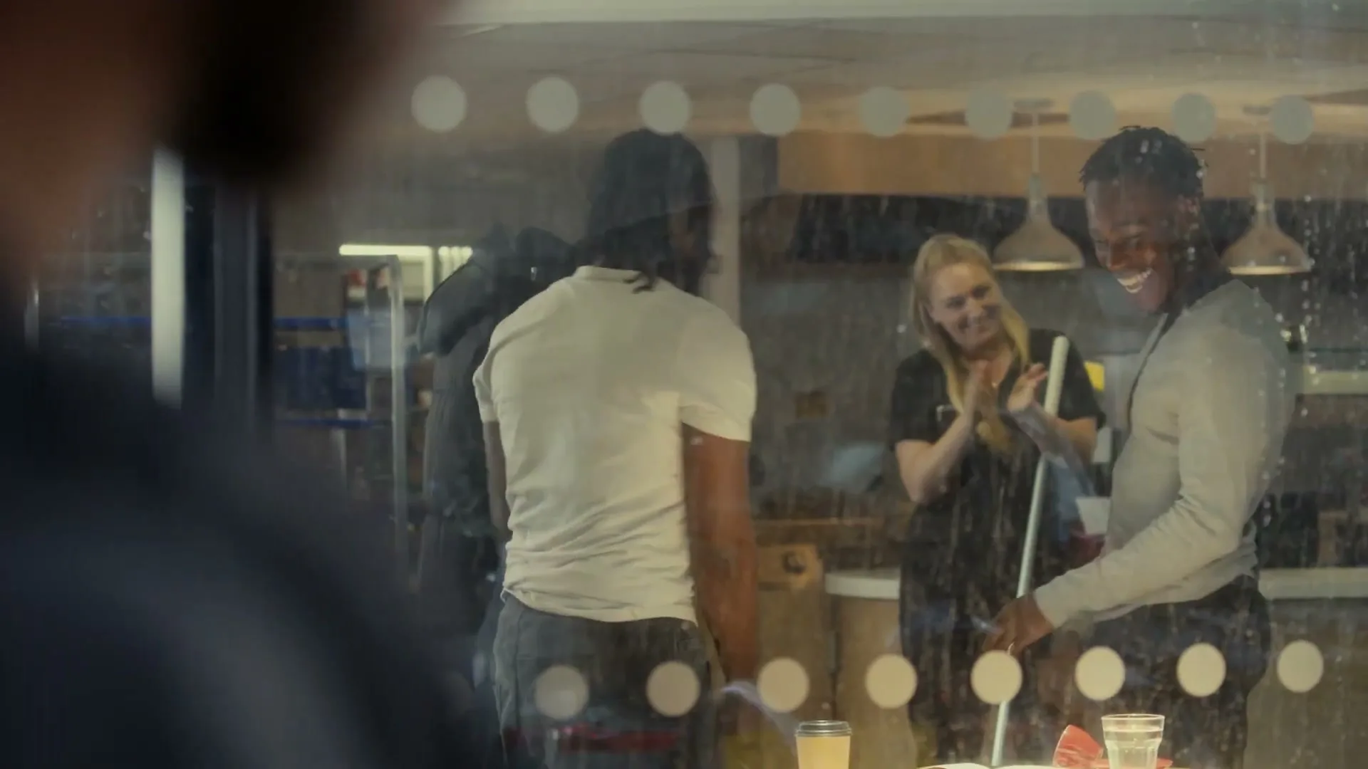 Two young men and a woman are seen through a window, with the woman clapping and smiling at the men, who are also smiling.