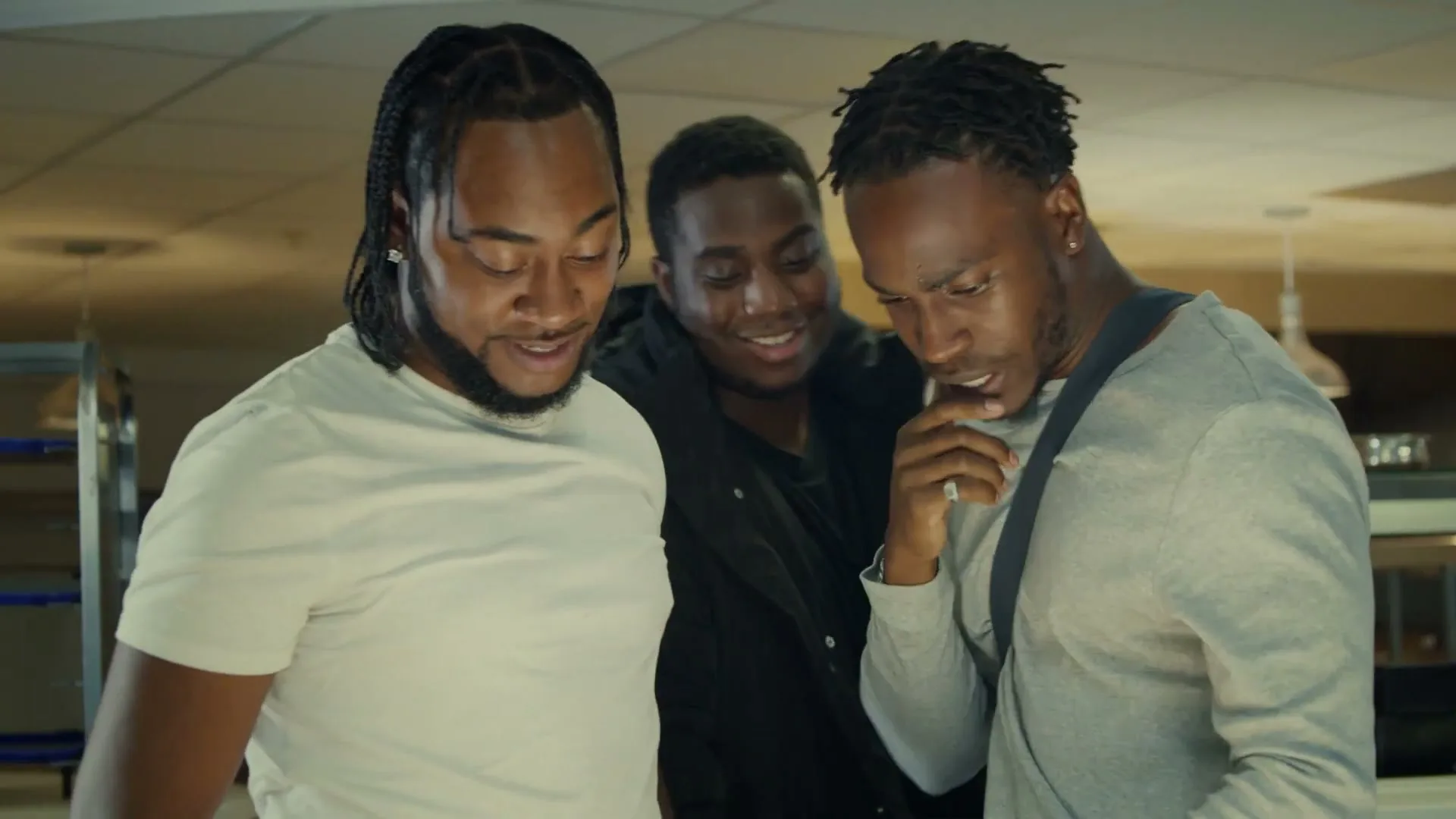 Three young Black men, two with braided hair, look down at something with varied expressions in a brightly lit interior space.
