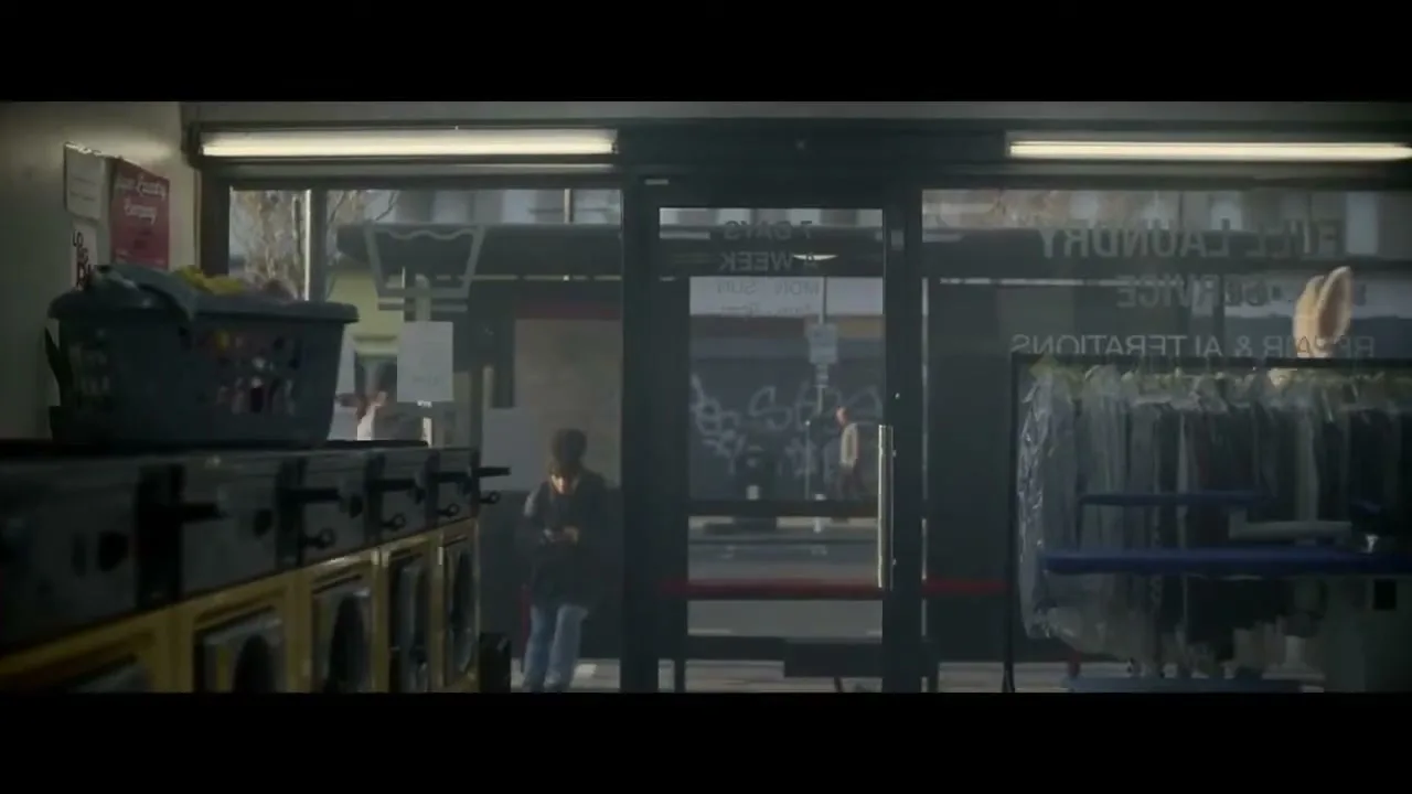 A person in a rabbit costume enters a laundromat, walking past washing machines and a rack of clothes, with another person visible outside.