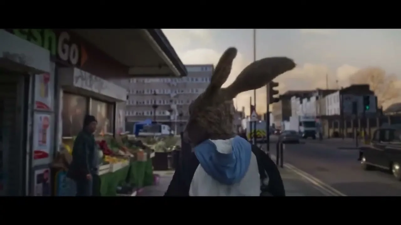 A person in a rabbit costume walks past a fruit stand on a city street, with buildings and cars in the background.