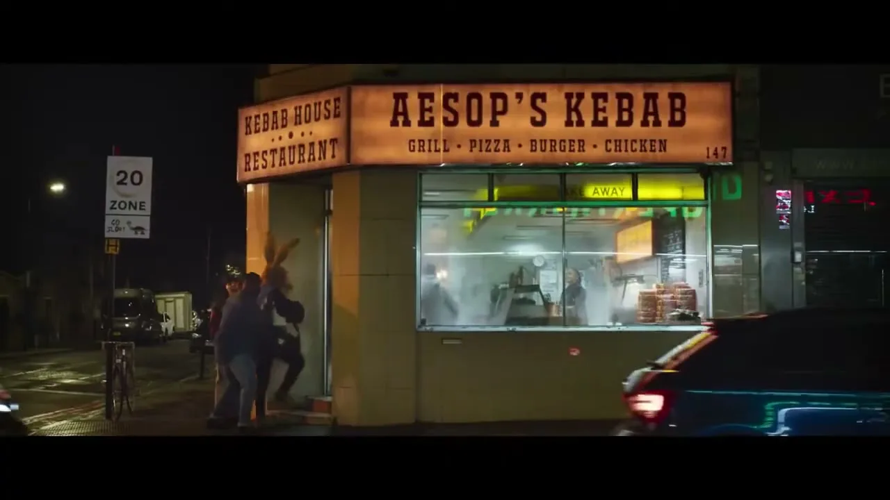 A group of people, including one in a hare costume, are seen outside "Aesop's Kebab" restaurant at night, as cars drive by.