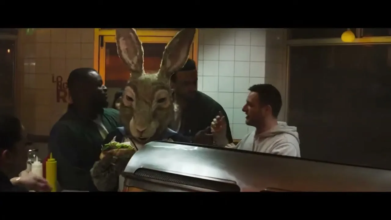 A person in a realistic hare costume receives a burger from a server at a busy fast-food counter, surrounded by other customers.
