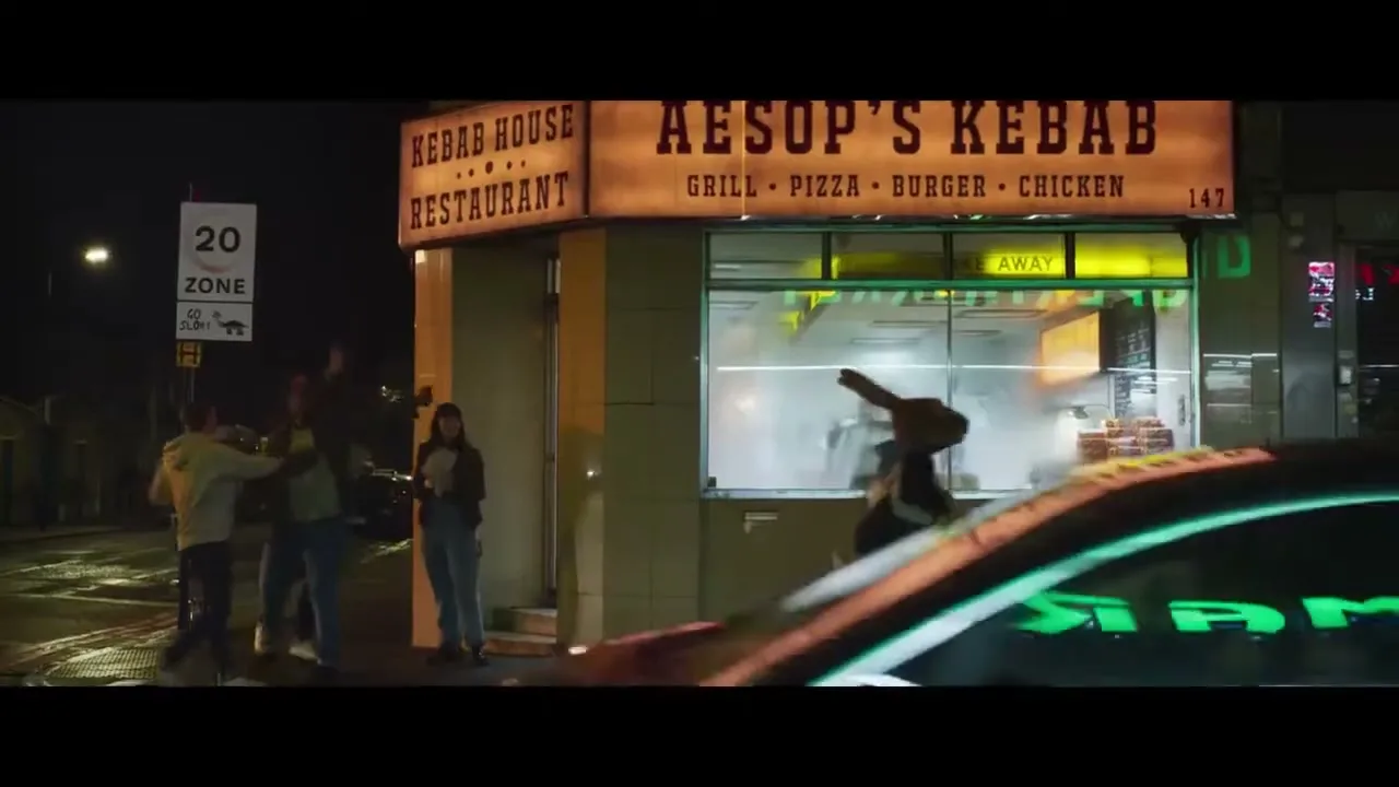 A person in a rabbit costume runs past a kebab shop at night, while a group of people stand outside on the wet pavement.