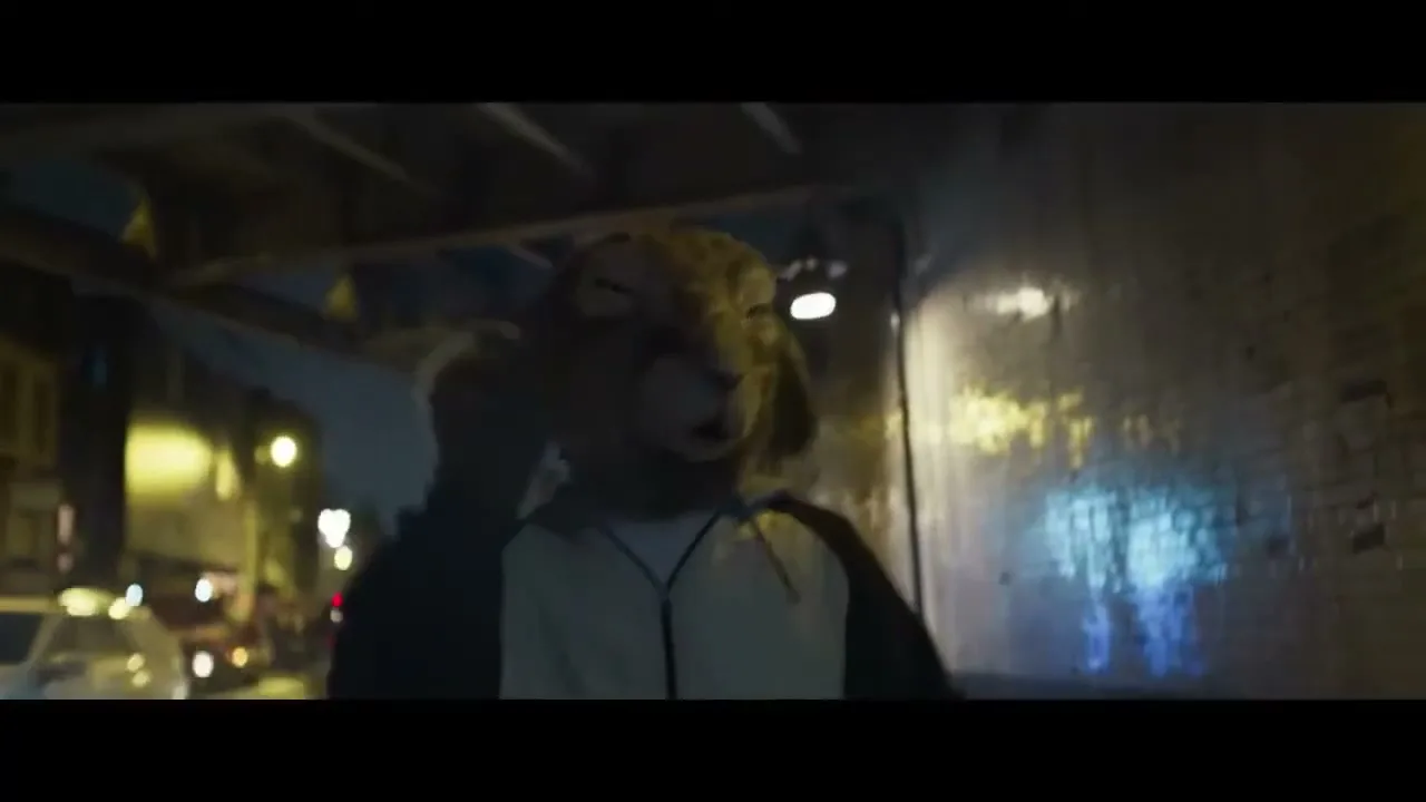 A person in a hare mask and hoodie walks along a dark city street at night, with cars and buildings visible in the background.