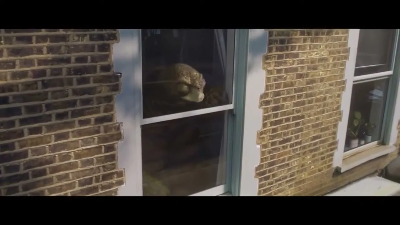 A large, brown, turtle-like creature with big eyes looks out of a window from inside a brick house.
