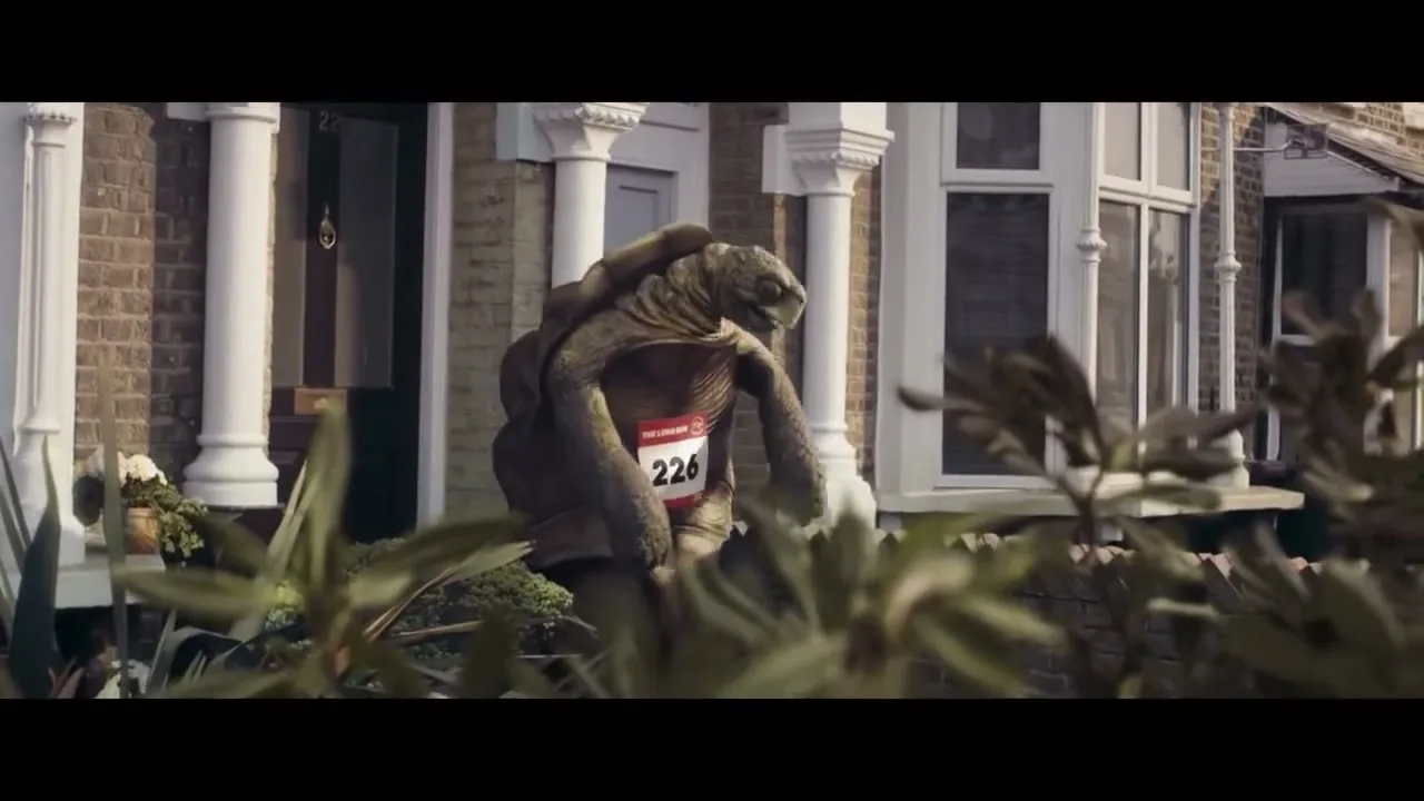 A person in a realistic tortoise costume, wearing a red race bib with "226" on it, slowly walks past a row of brick houses on a sunny day.