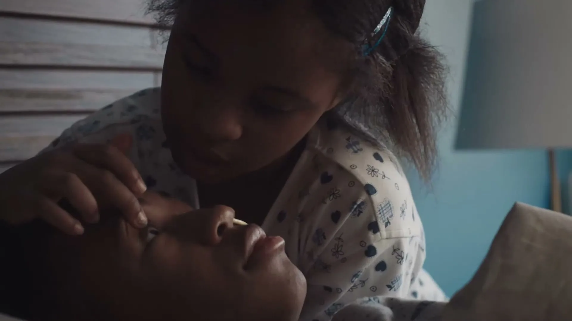 A young girl with dark hair and a patterned shirt gently touches the face of a sleeping adult, possibly her mother, in a dimly lit bedroom.